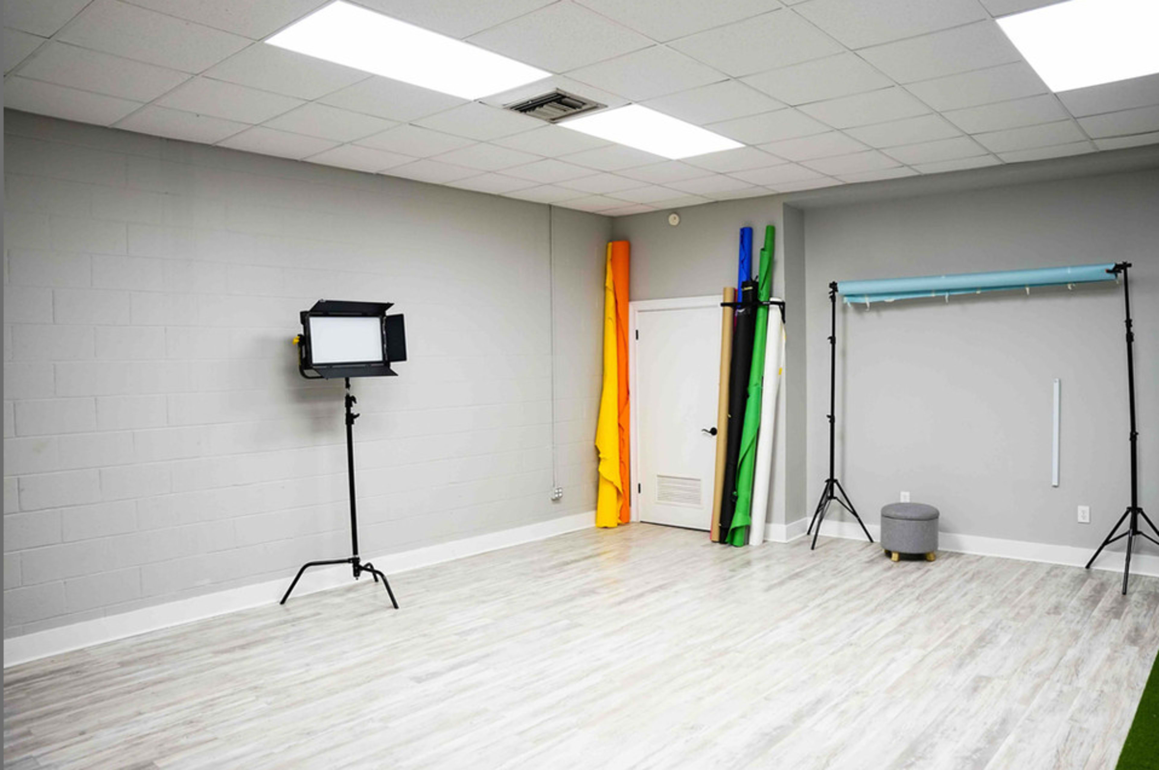 A spacious studio with gray walls, a light wooden floor, a lighting setup on a tripod, and colorful backdrops leaning against the wall.