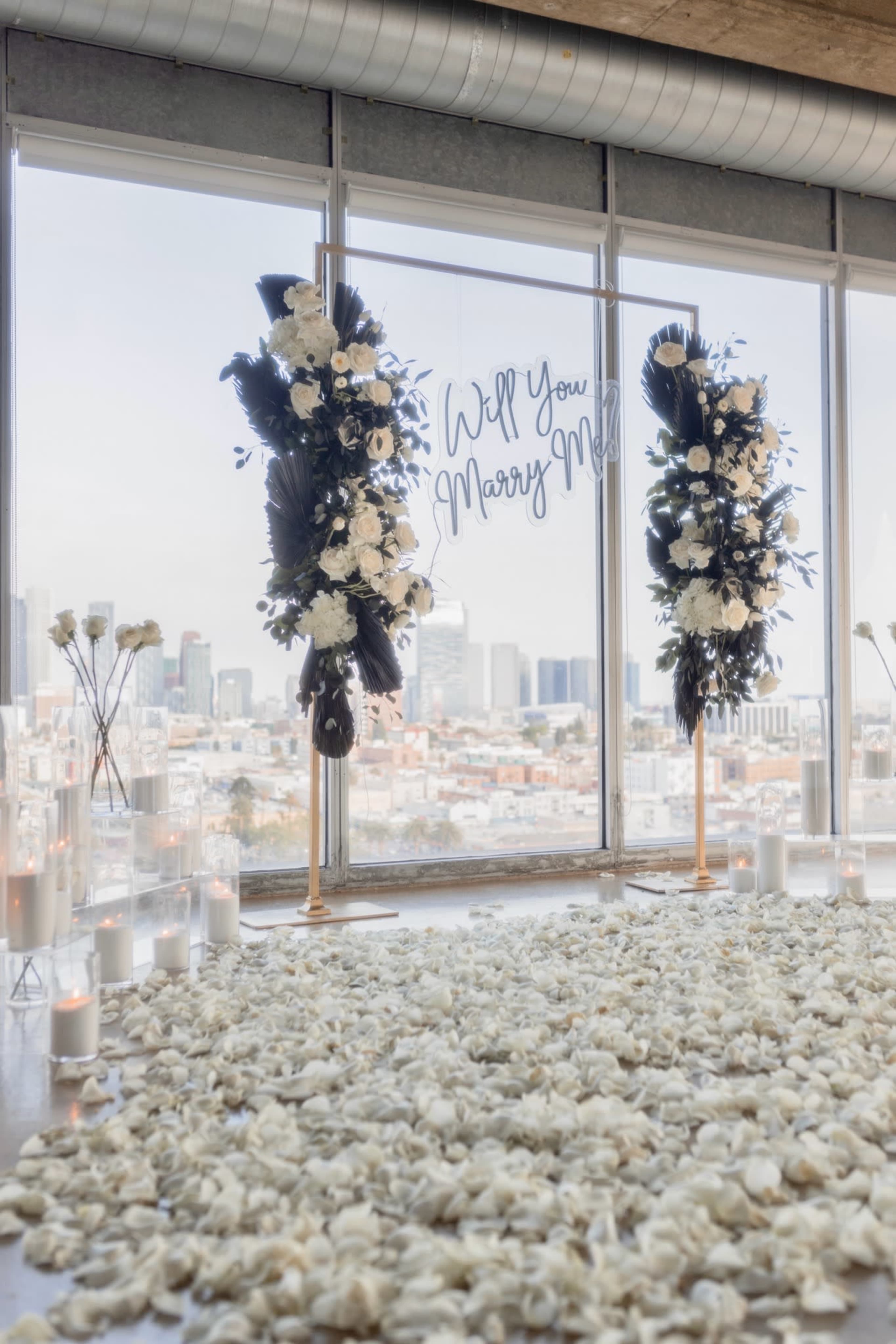 A romantic proposal setup features a large floral display on either side of a glass window with a view of the city, complemented by a carpet of rose petals and candles.