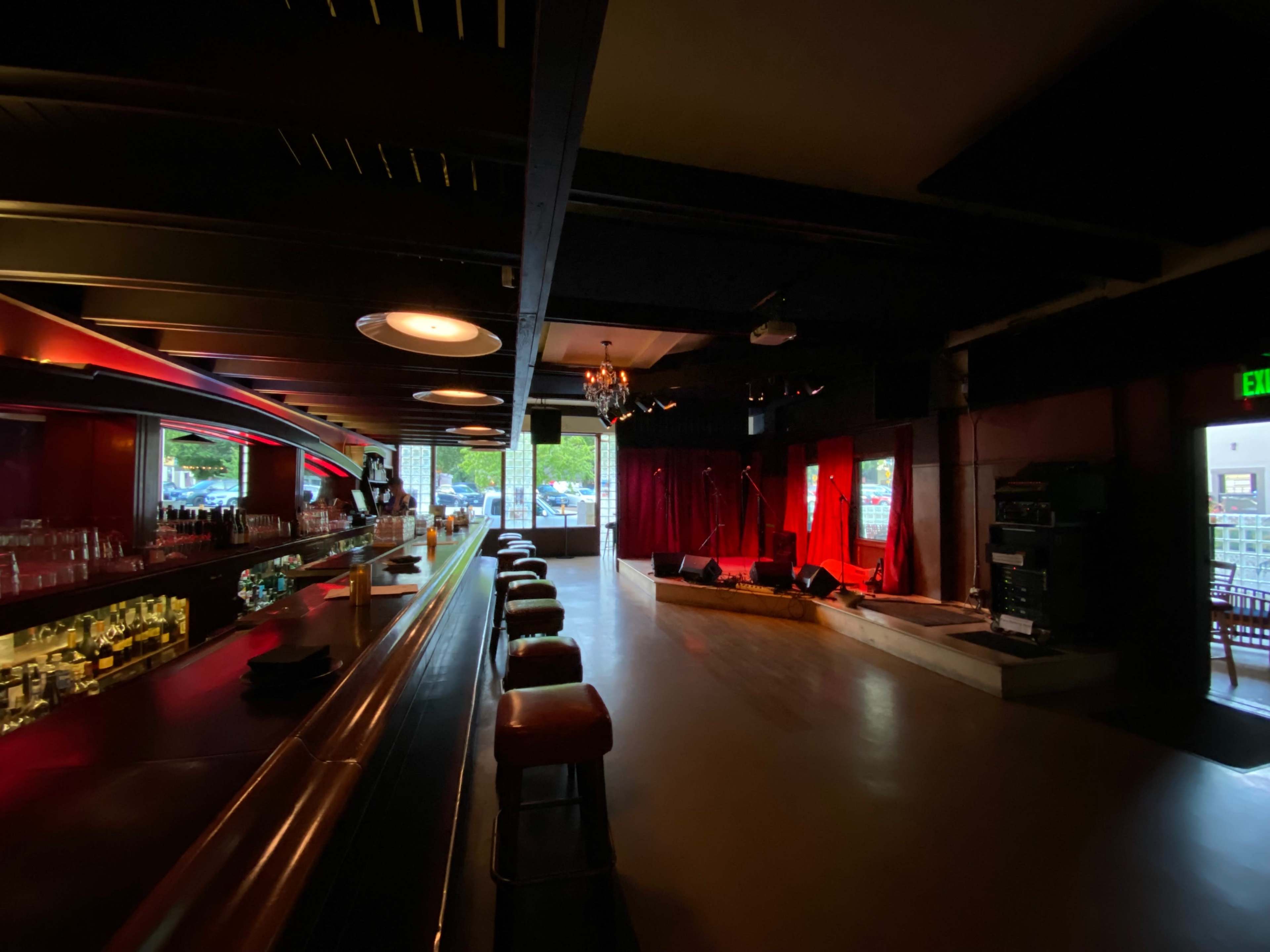 A dimly lit bar with a long counter and several stools facing a small stage with red curtains, illuminated by soft lighting.