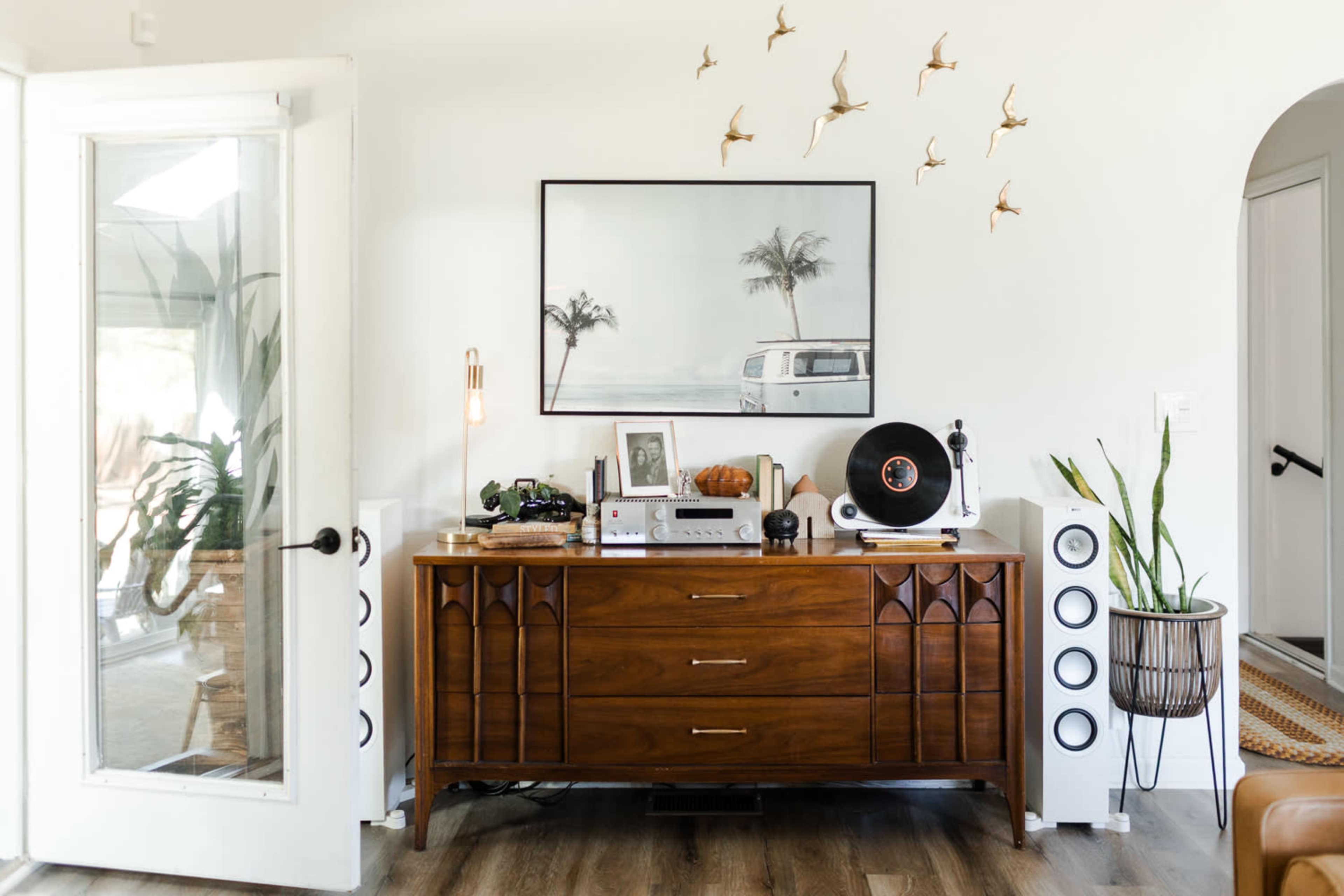 A wooden dresser is positioned against a white wall, decorated with a framed photograph and small bird sculptures, with a turntable and speakers on either side.
