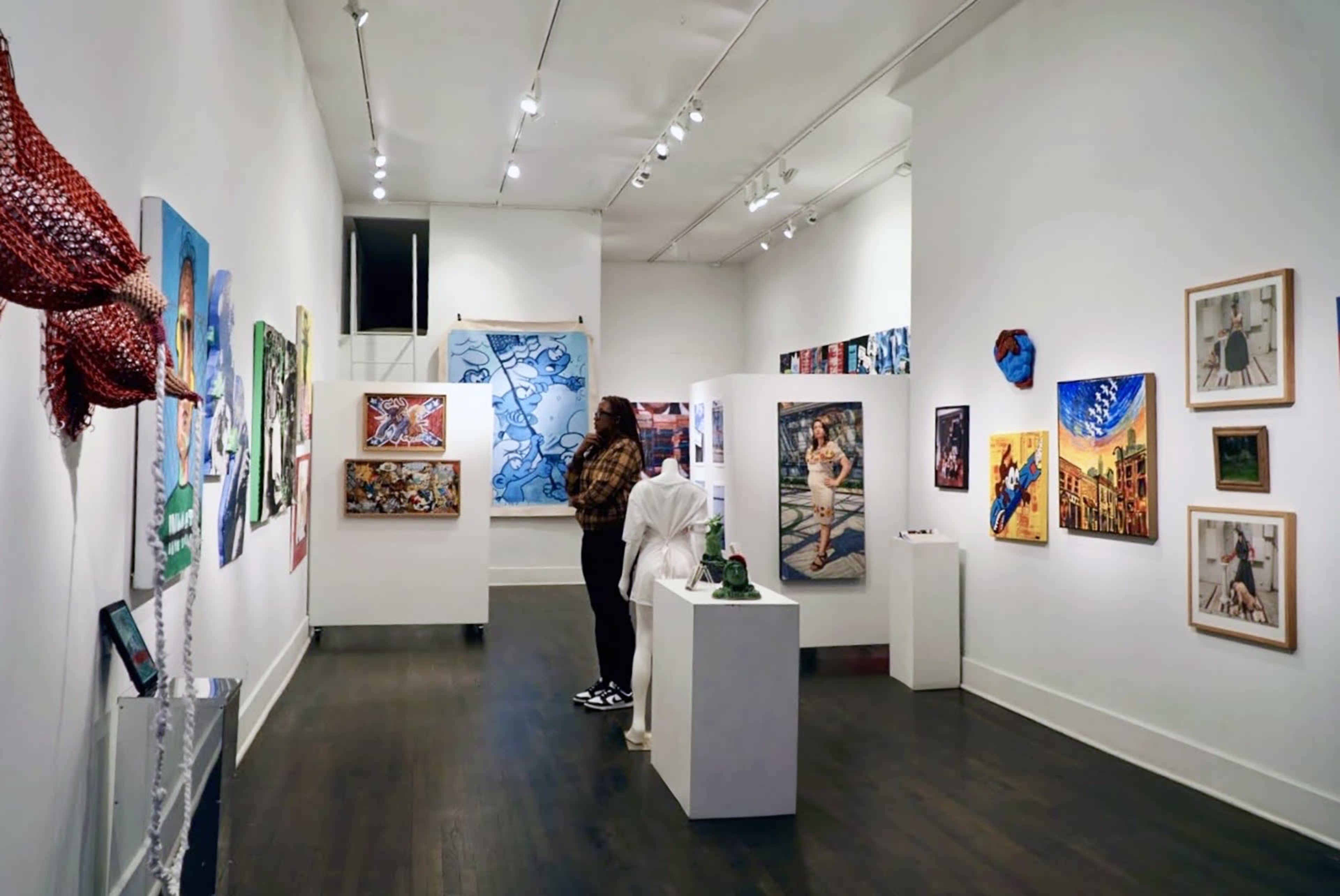 A person stands in an art gallery, examining various colorful paintings displayed on the walls and on pedestals.