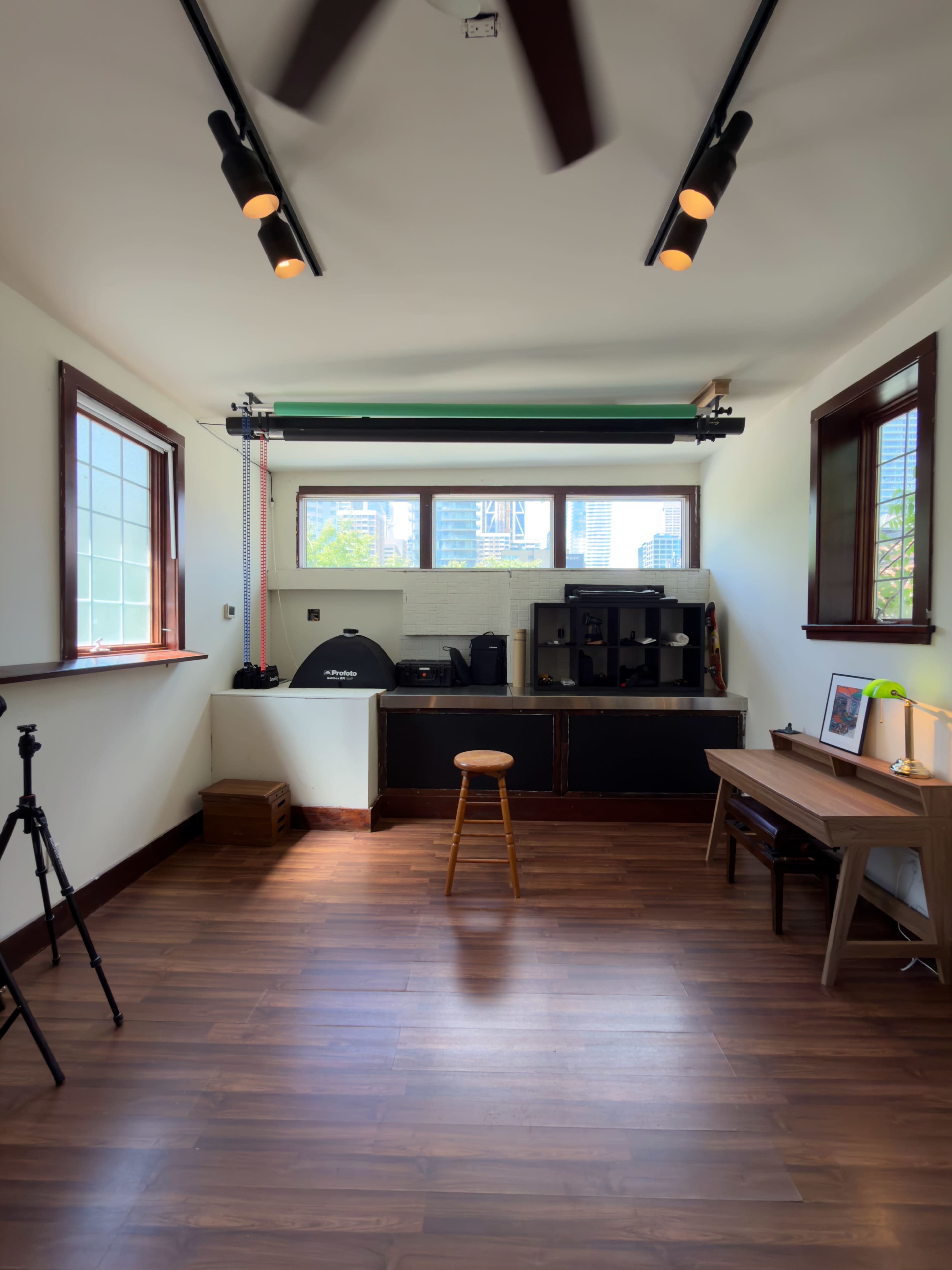The image shows a minimalist room with wooden flooring, a stool in the center, a desk on the right, and a window with city views in the background.