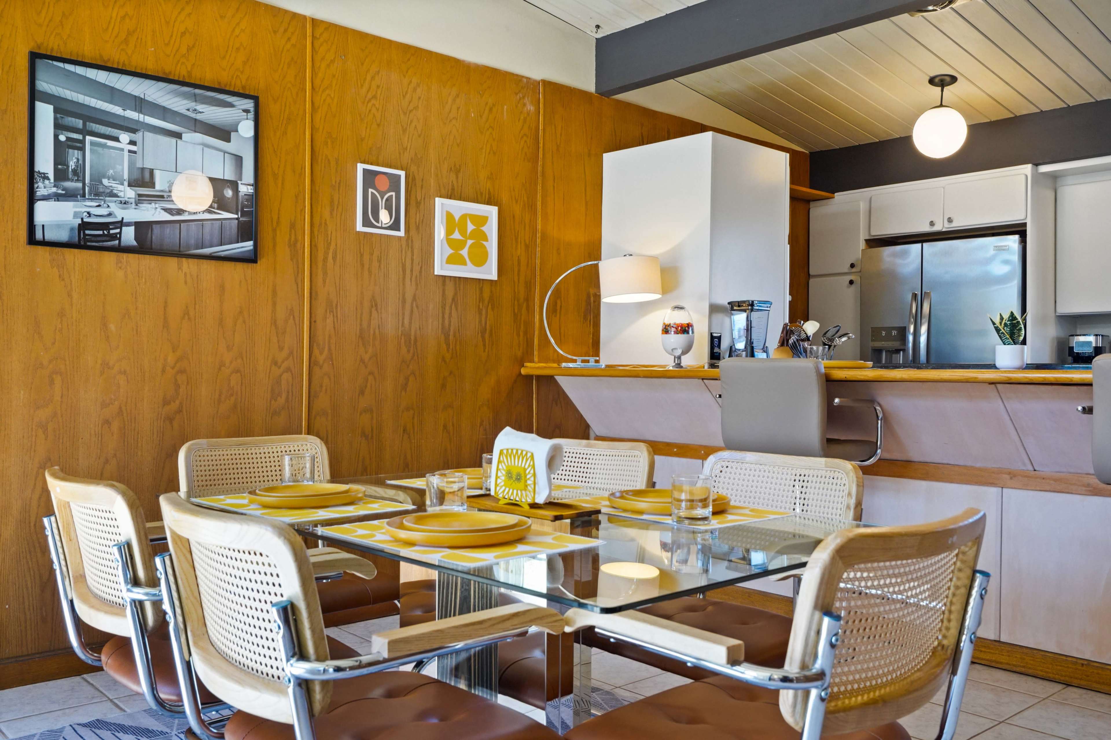 A dining area features a glass table surrounded by chairs, with wood-paneled walls and a kitchenette in the background.
