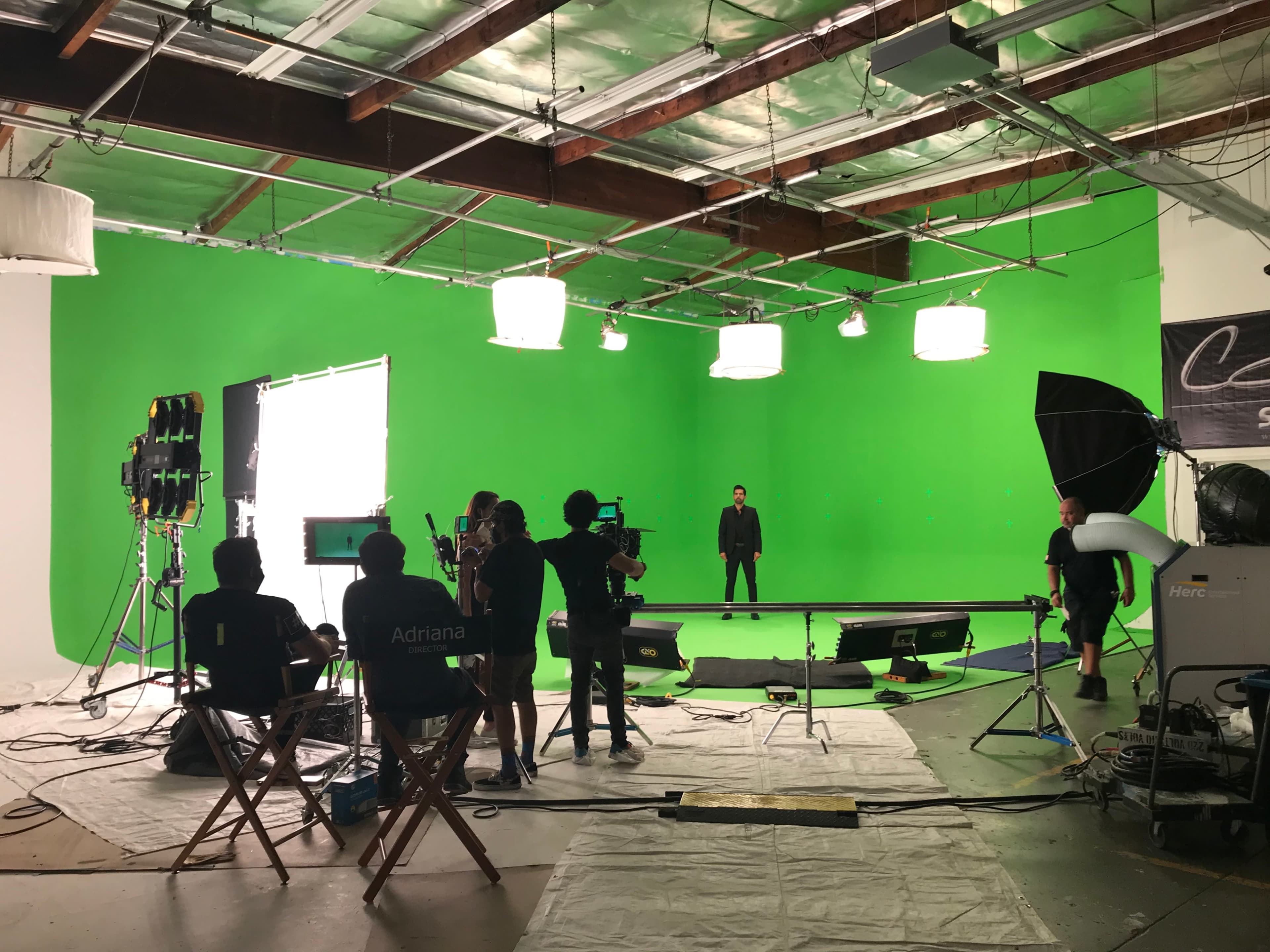 A film crew works on a green screen set with a lighting setup and several cameras aimed at a performer in the center of the studio.