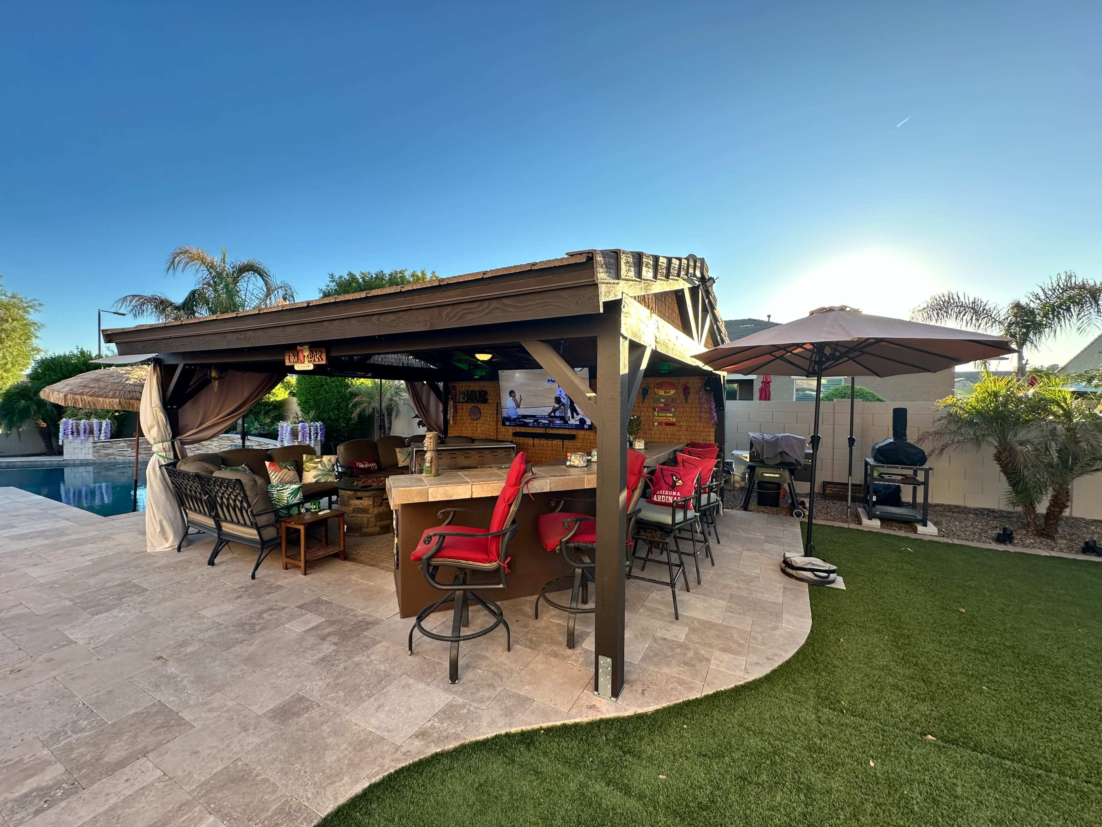 A covered outdoor space features a bar area with red chairs, nearby seating, and a pool in the background.