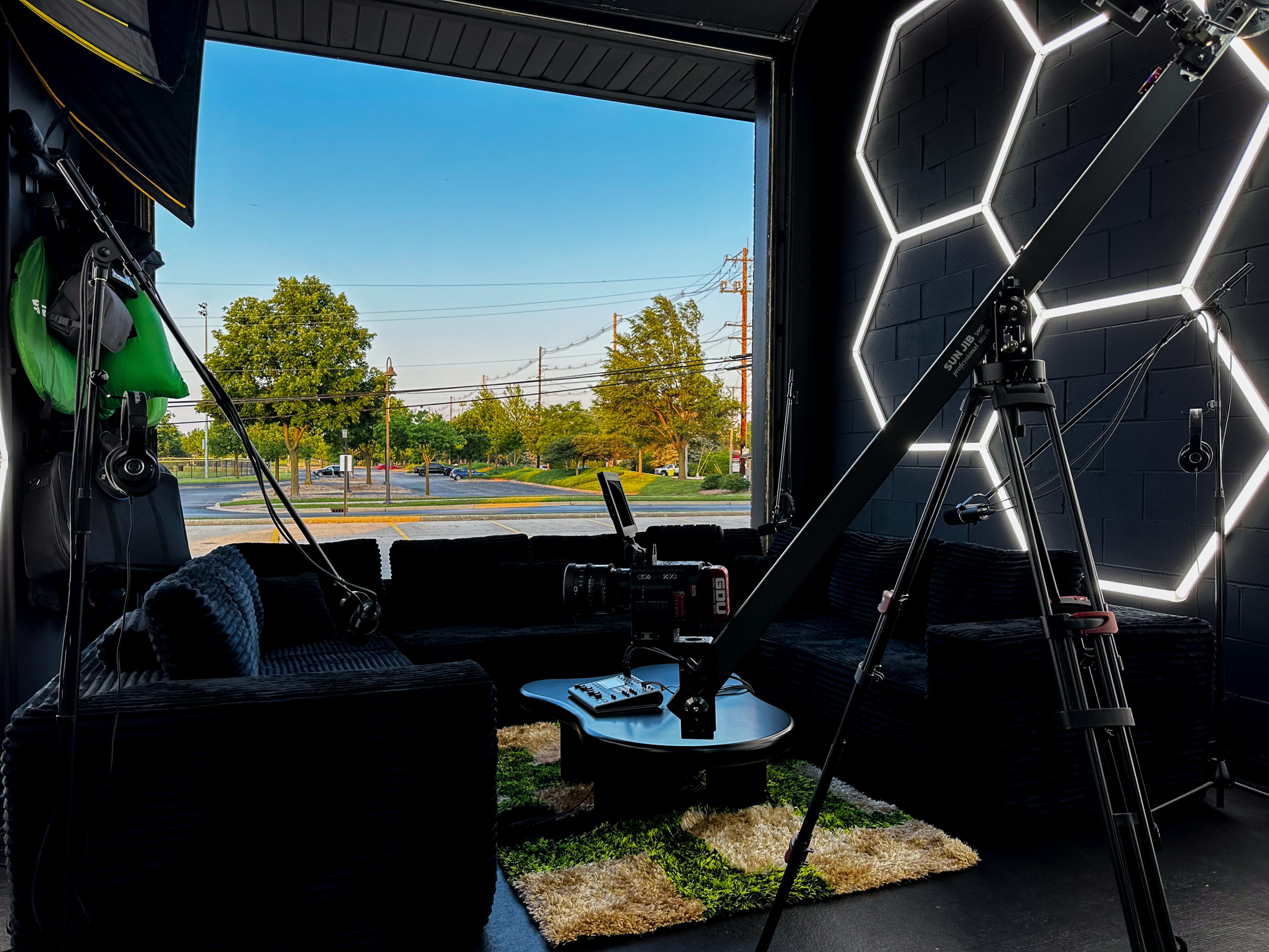 The scene shows a modern studio setup with black furniture, a camera on a tripod, and a view of an open entrance featuring greenery and power lines outside.