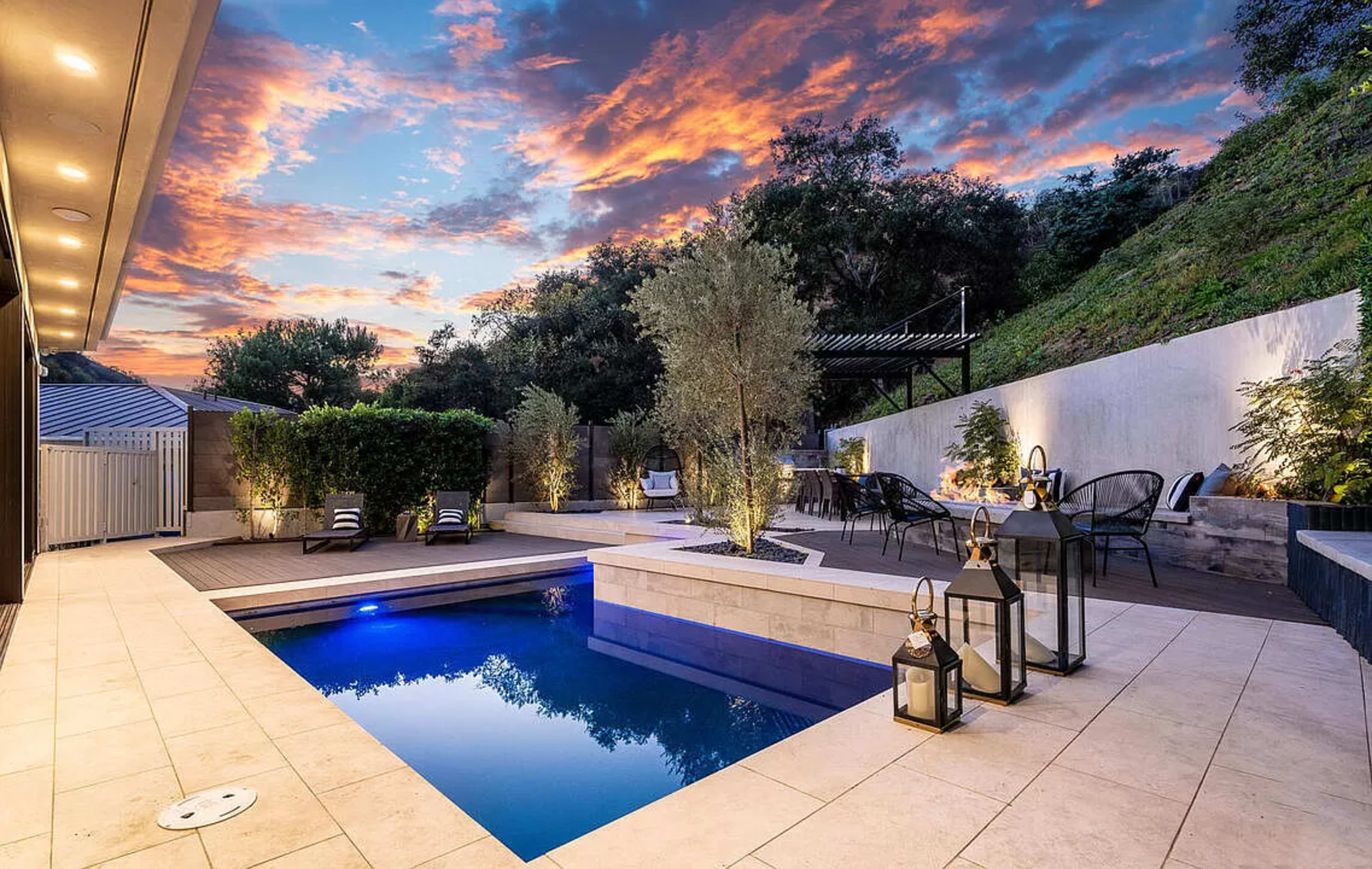The image shows a modern outdoor space featuring a small swimming pool surrounded by landscaped greenery and seating areas, set against a colorful sunset sky.
