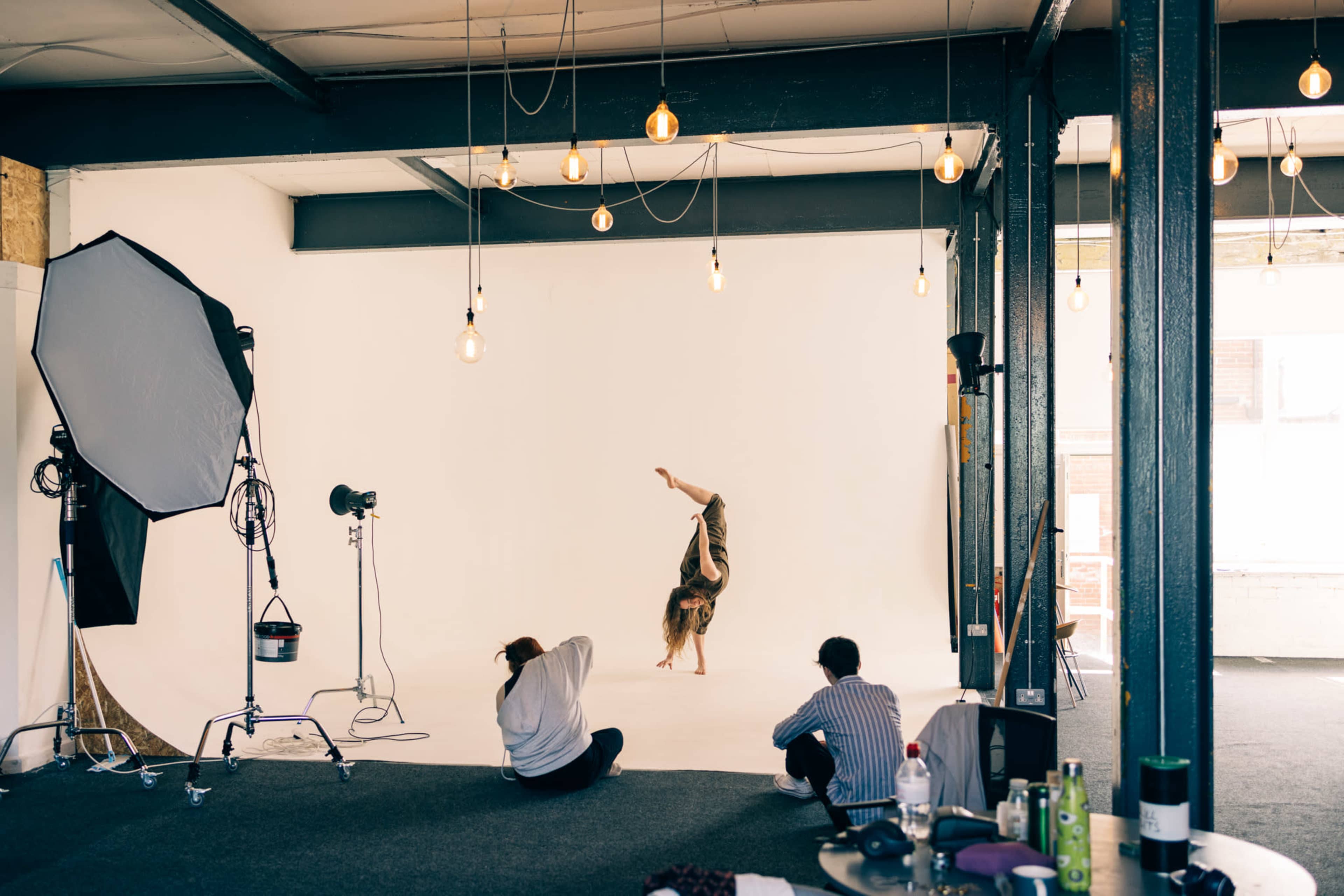 A person performs a handstand in a photography studio while two onlookers observe.