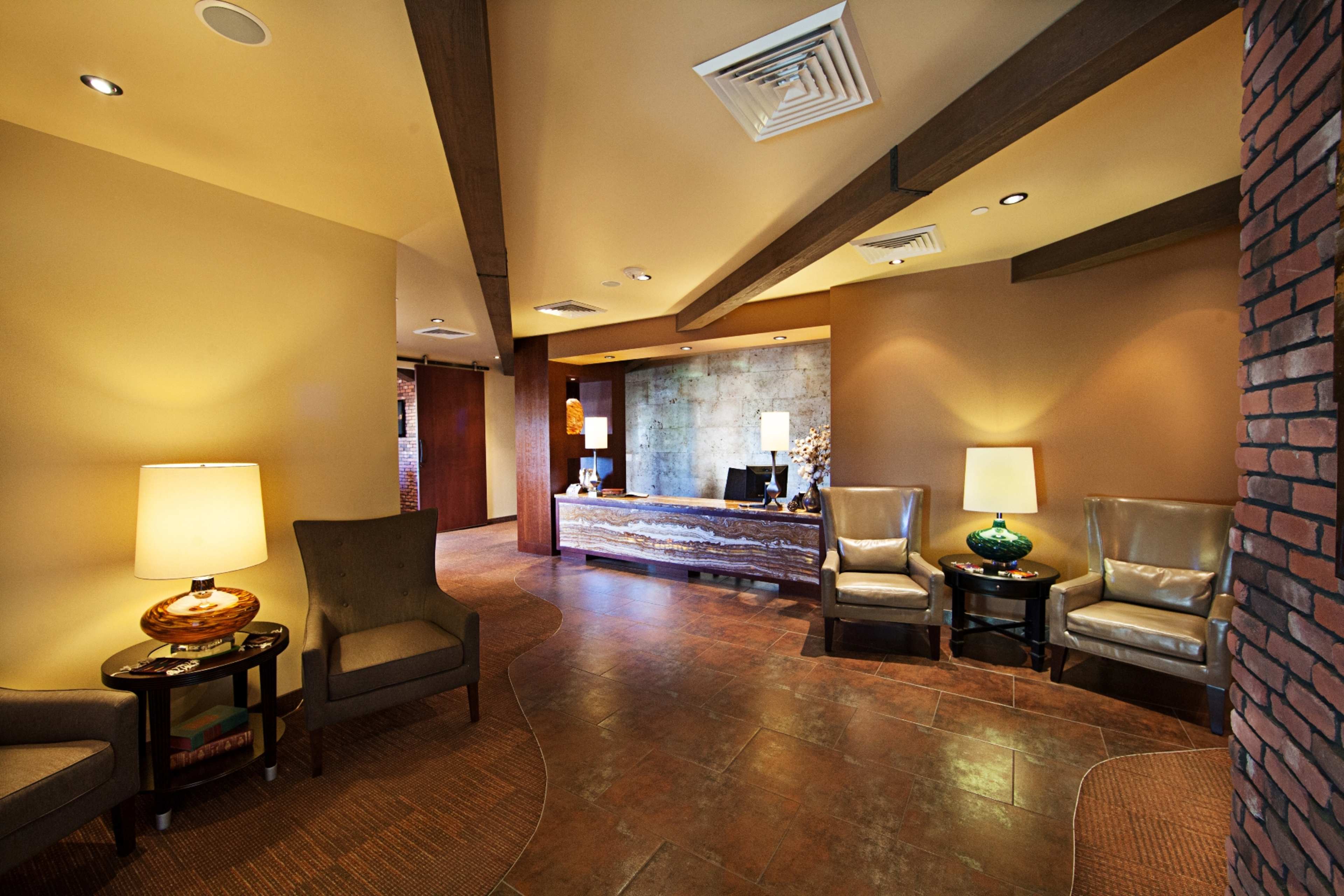 A lobby area with two armchairs, a side table, a reception desk, and decorative lamps against a warm-toned wall.