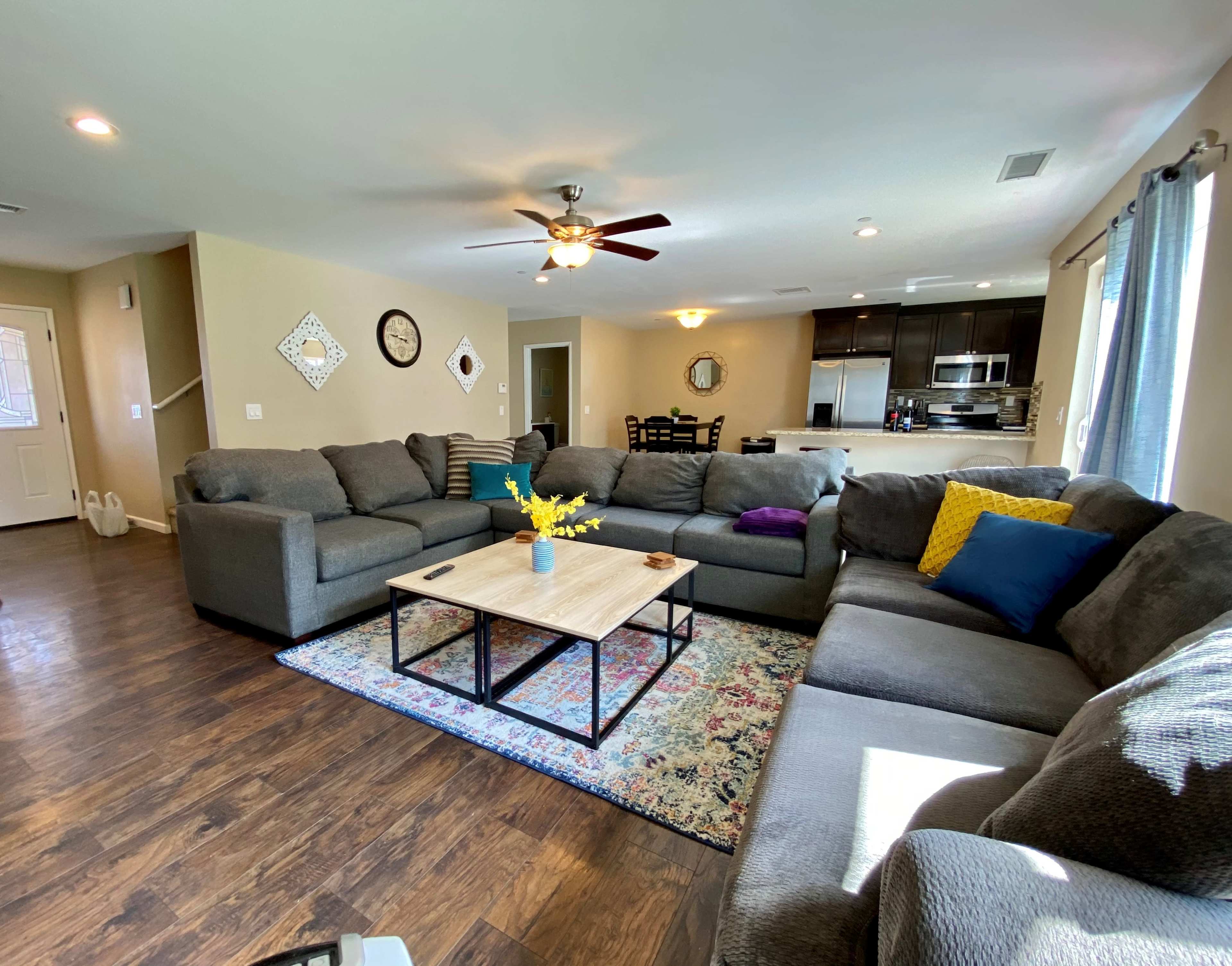 The living room features a large sectional sofa arranged around a coffee table, with a colorful area rug and a view into the kitchen and dining area.