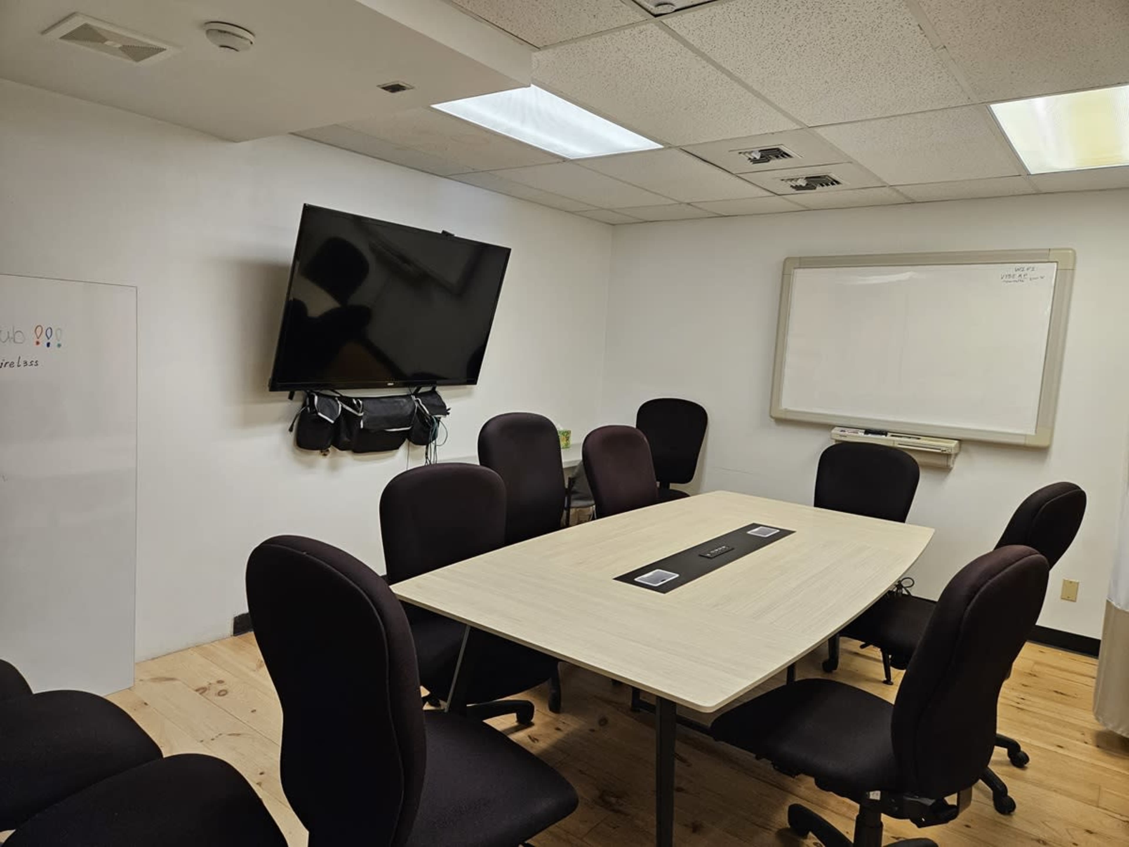 A conference room features a large table surrounded by eight black chairs, a wall-mounted TV, and a whiteboard.