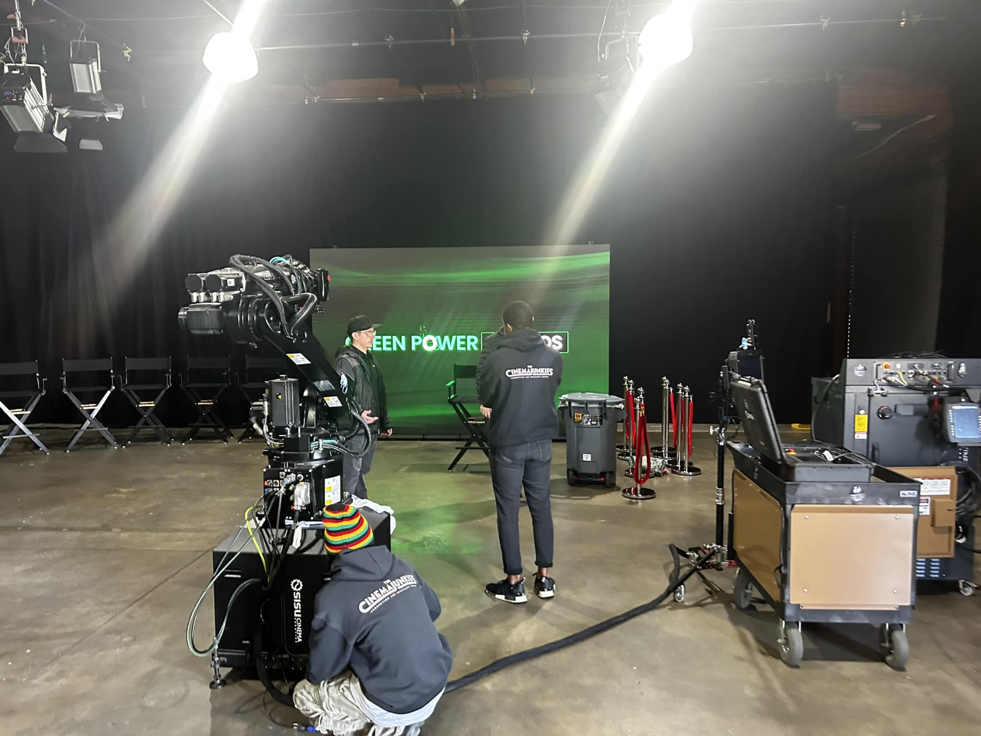 A television production studio is set up with a camera, crew members, and a backdrop displaying "GREEN POWERED."