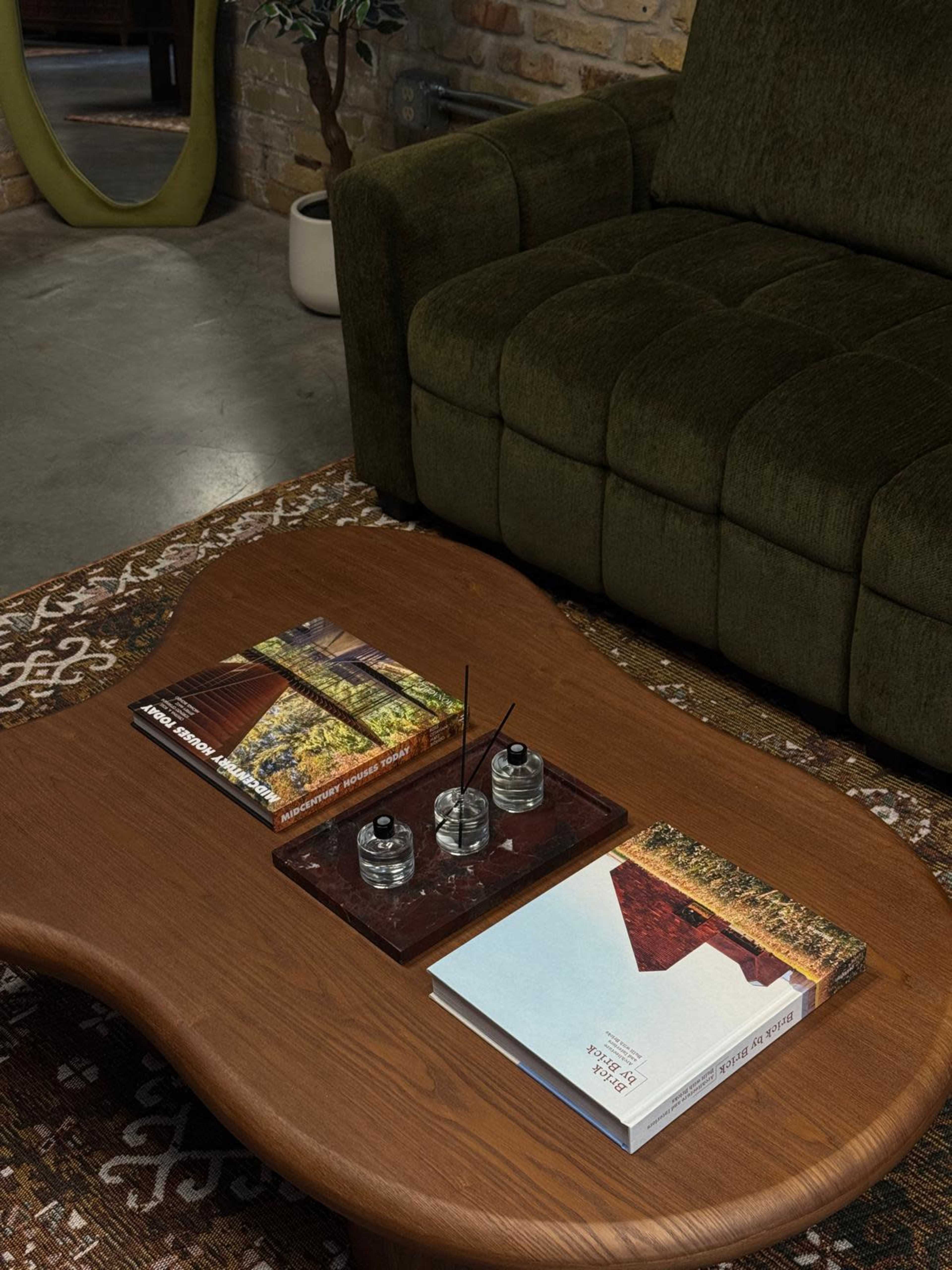 A wooden coffee table with a curved shape holds two books and a tray of scented candles, positioned in front of a green upholstered couch in a cozy interior space.