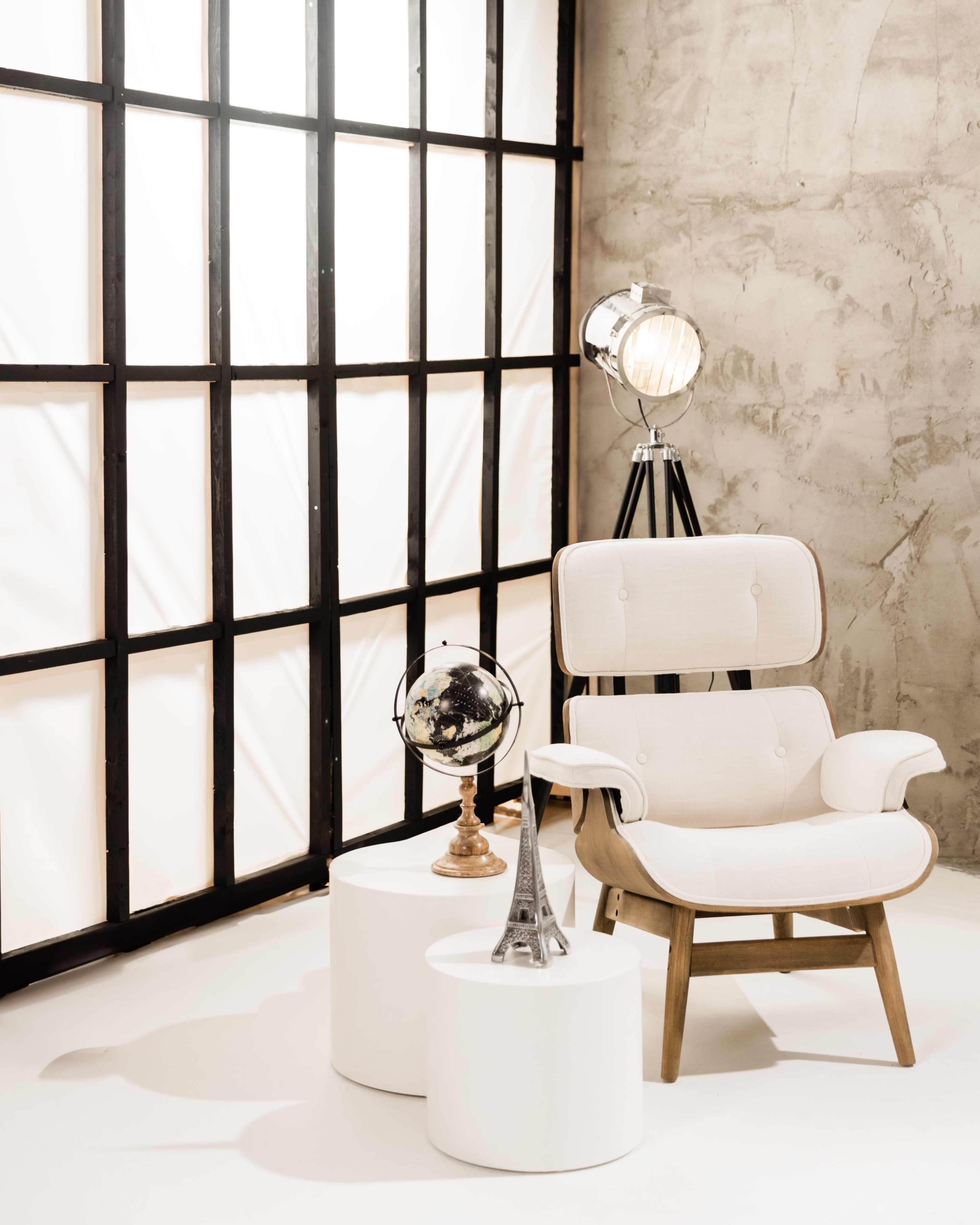 The scene features a minimalist interior with a white chair next to two round tables, a globe, and a small Eiffel Tower replica against a backdrop of a large, grid-patterned window.