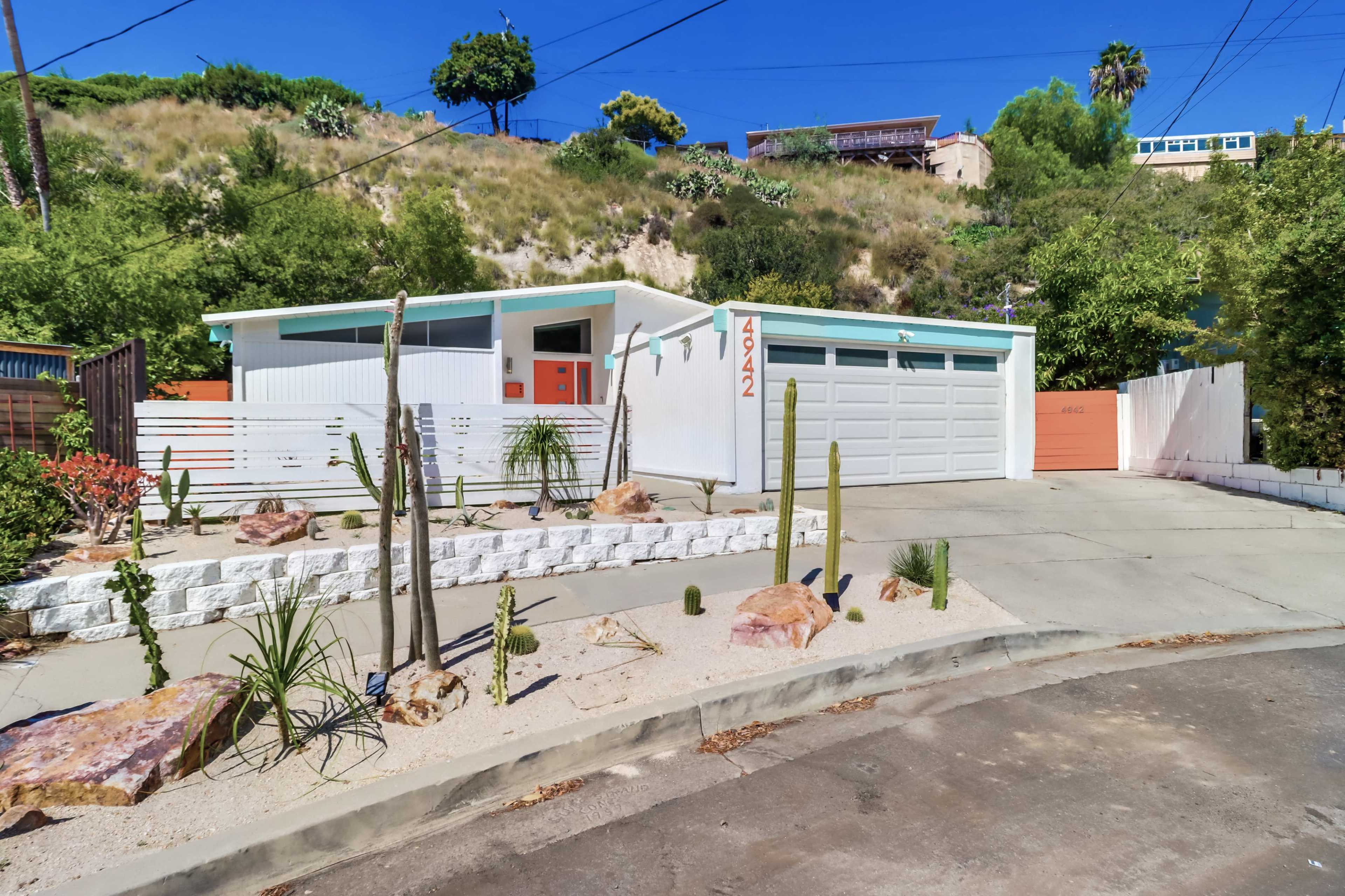 A modern house with a bright orange door, a two-car garage, and landscaping that includes cacti and rocks on a sloped street.