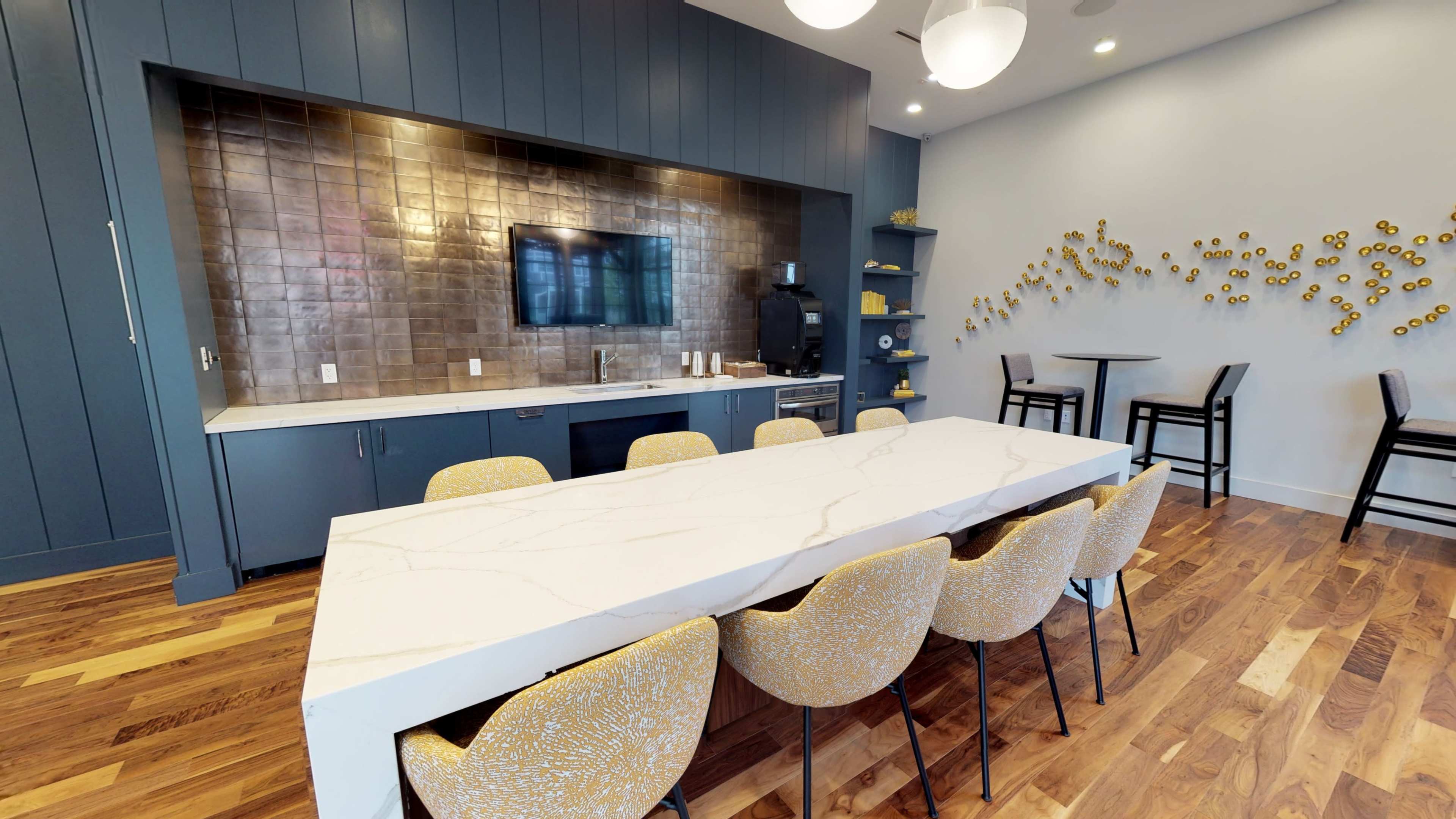 A modern kitchen area with a large marbled table and stylish seating, a backsplash of brown tiles, and a television mounted on the wall.