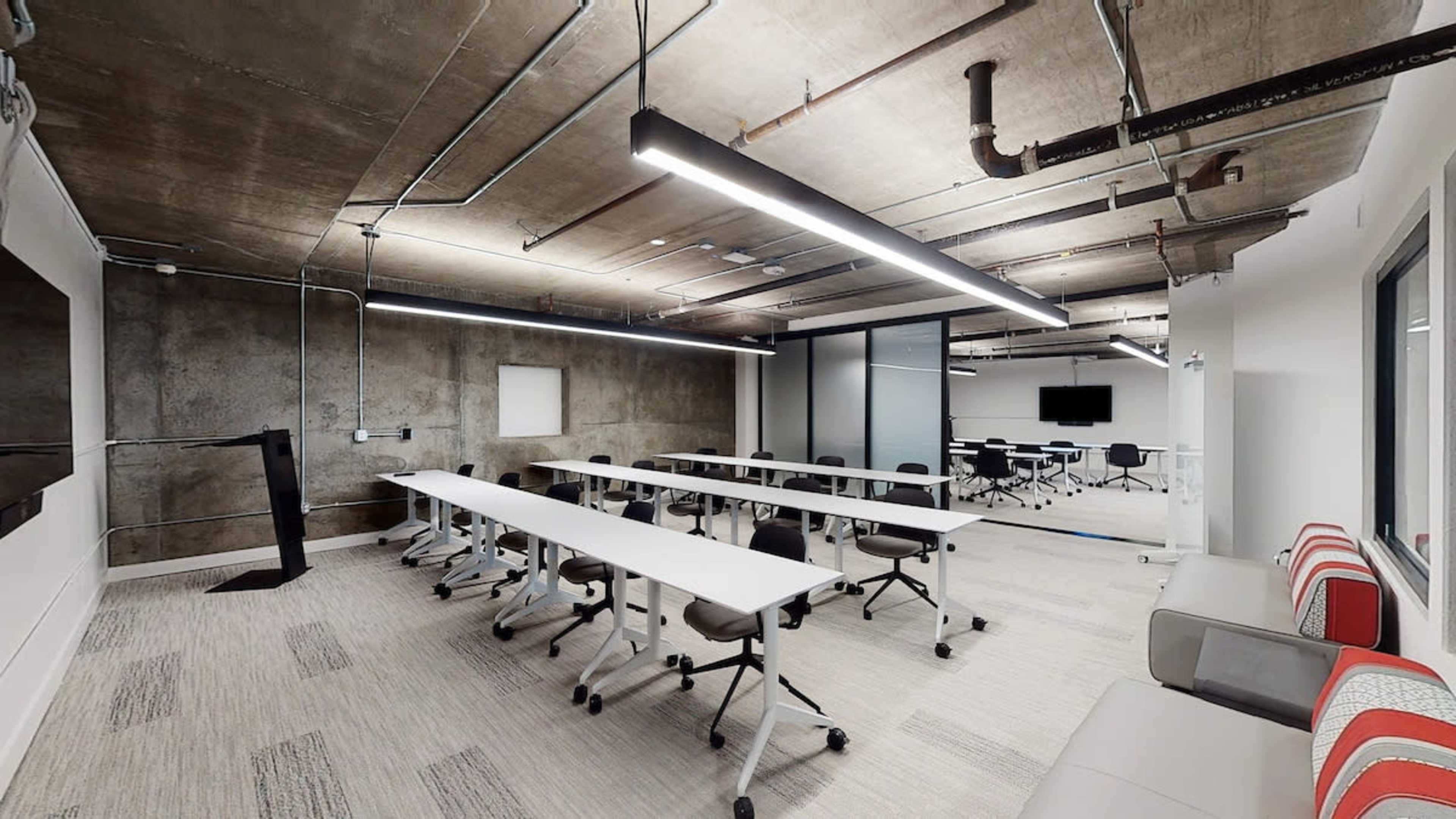 A modern conference room with rows of tables, chairs, and overhead lighting, featuring a concrete ceiling and large windows.