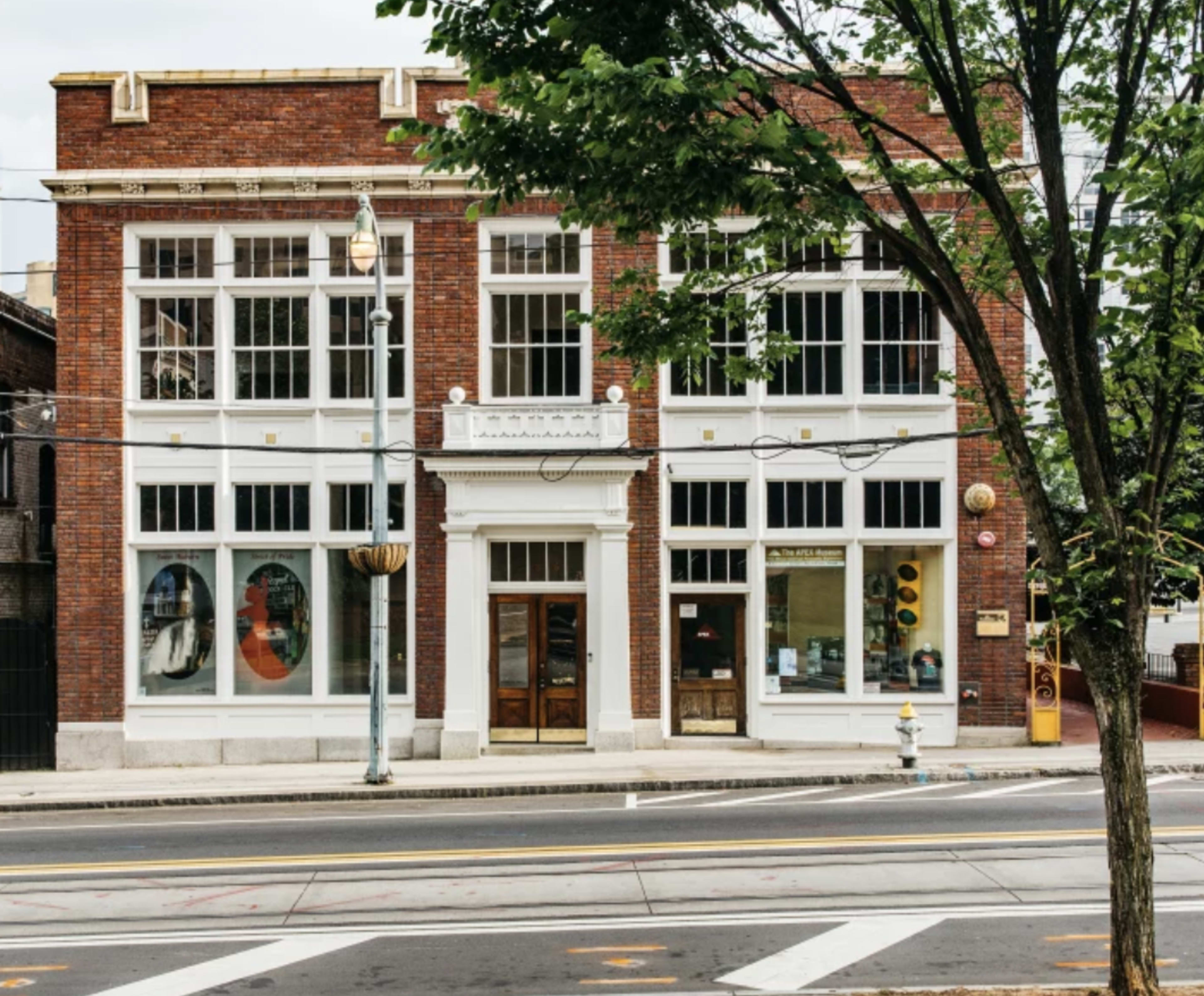 A two-story brick building with large windows and a decorative entrance, located on a street lined with trees.