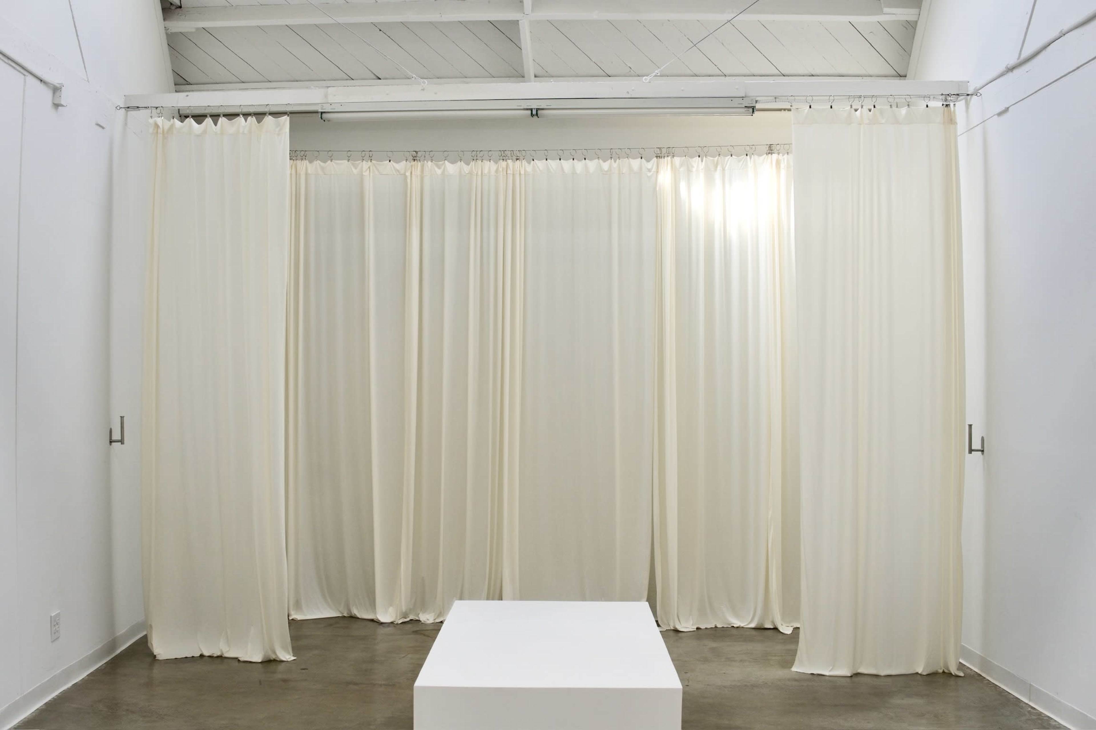 The image shows a minimalist interior space with white walls, a polished concrete floor, and sheer curtains dividing the area, along with a central white square pedestal.