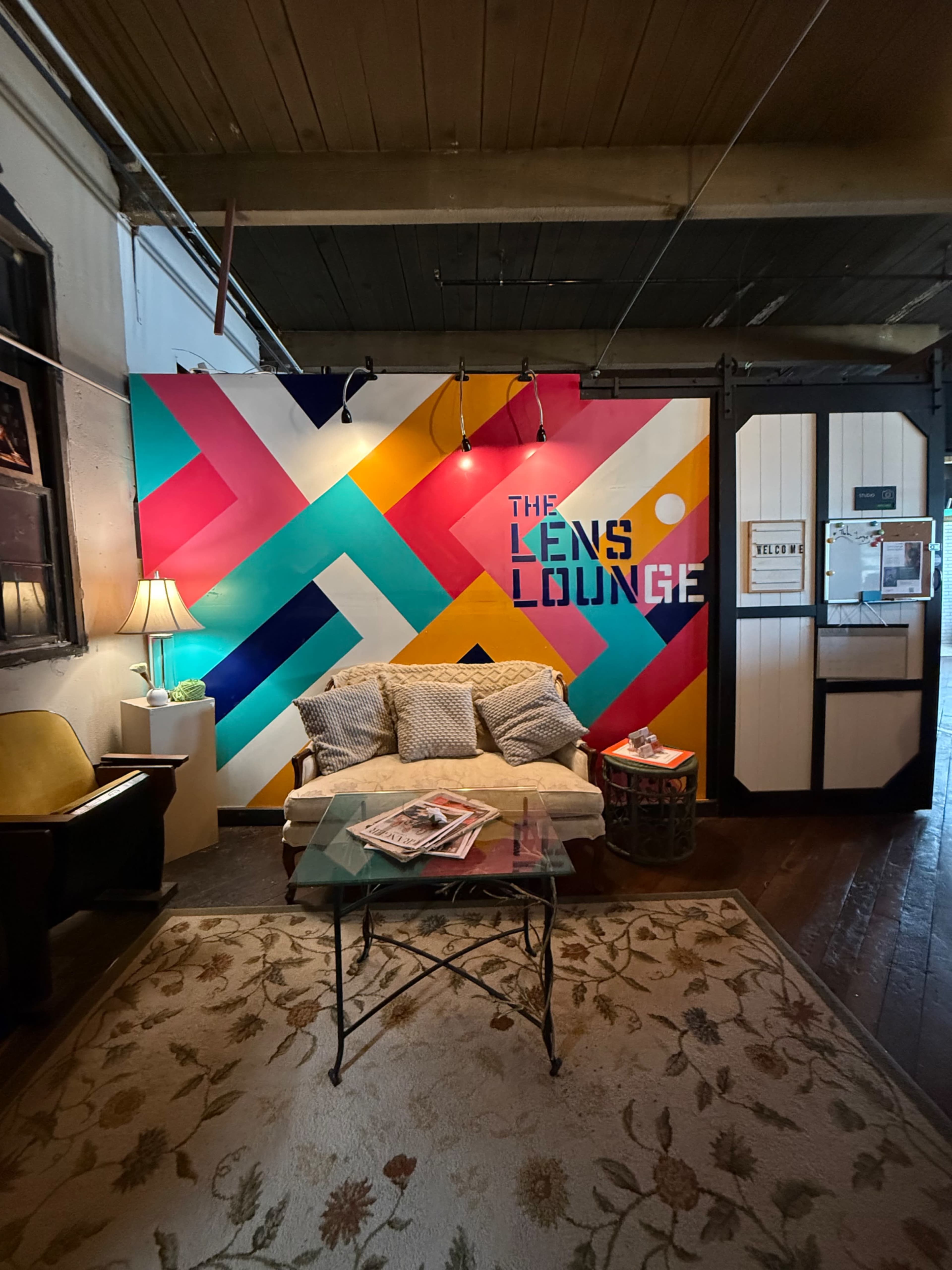 The image shows a cozy seating area in a space named "The Lens Lounge," featuring a vibrant, geometric mural and a glass coffee table.