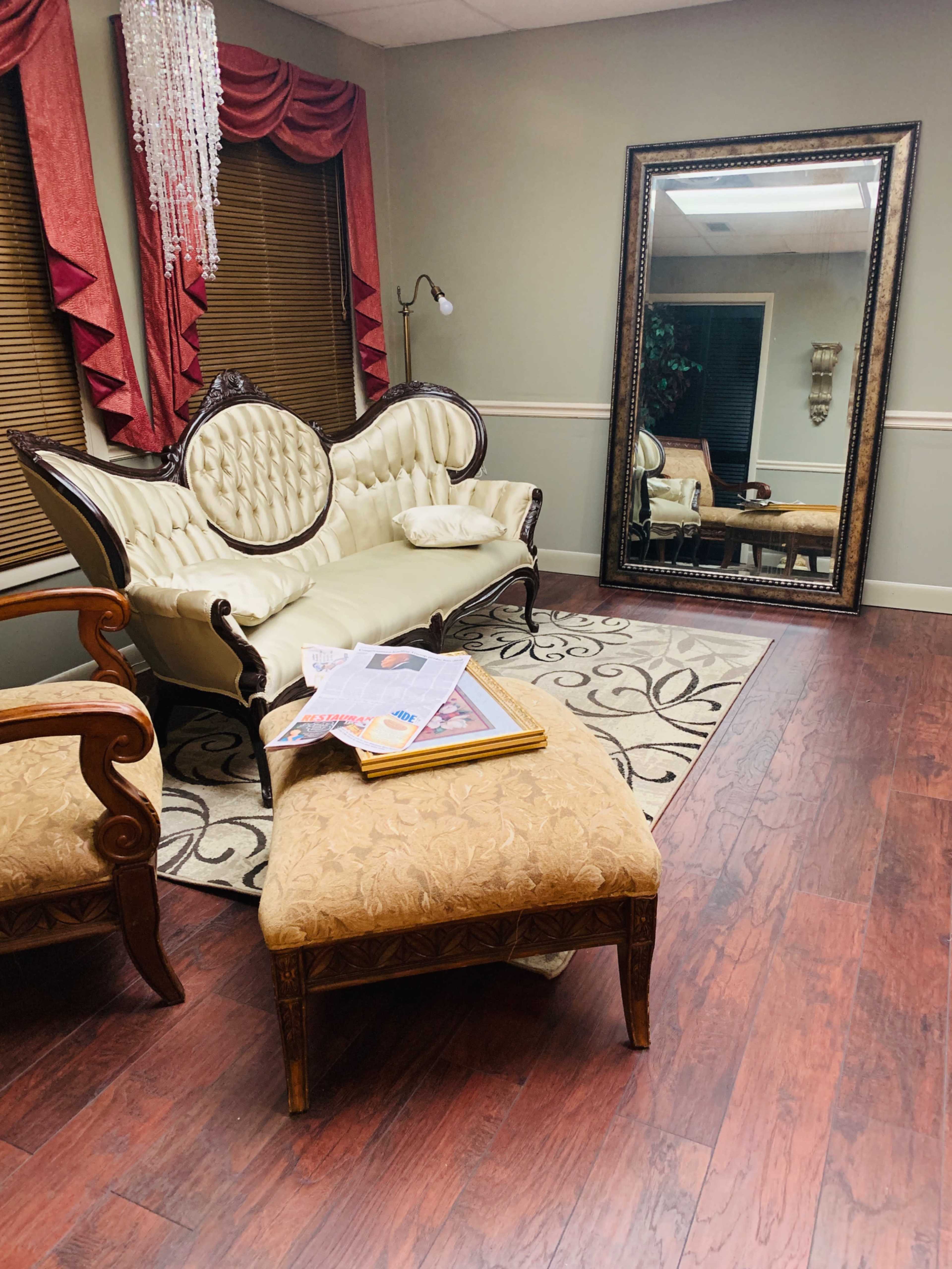 A furnished room with a vintage couch, an ottoman, and a large mirror, featuring a patterned rug and wooden flooring.
