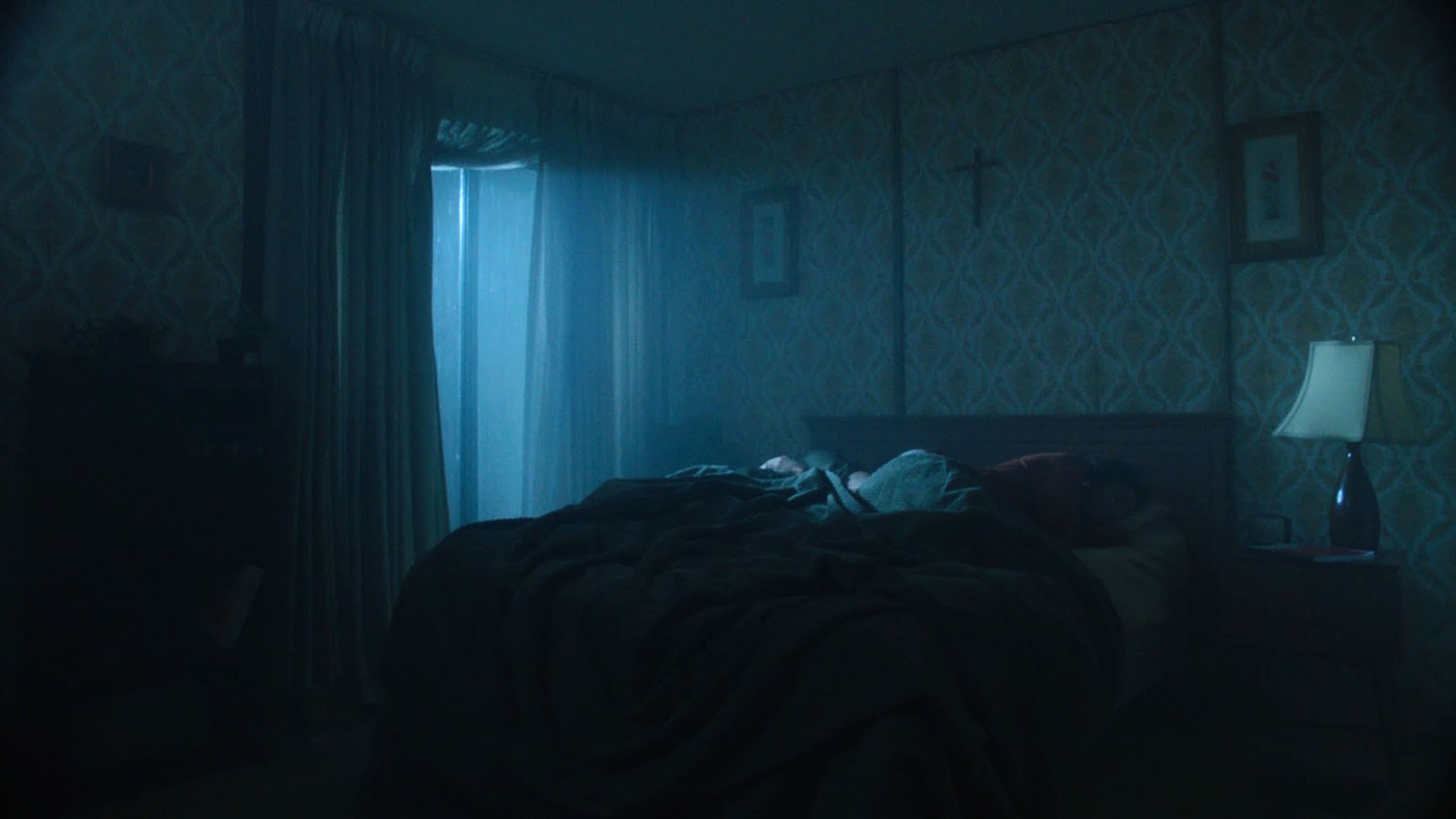A dimly lit bedroom features a person sleeping in a bed, with a window casting a beam of light into the room.