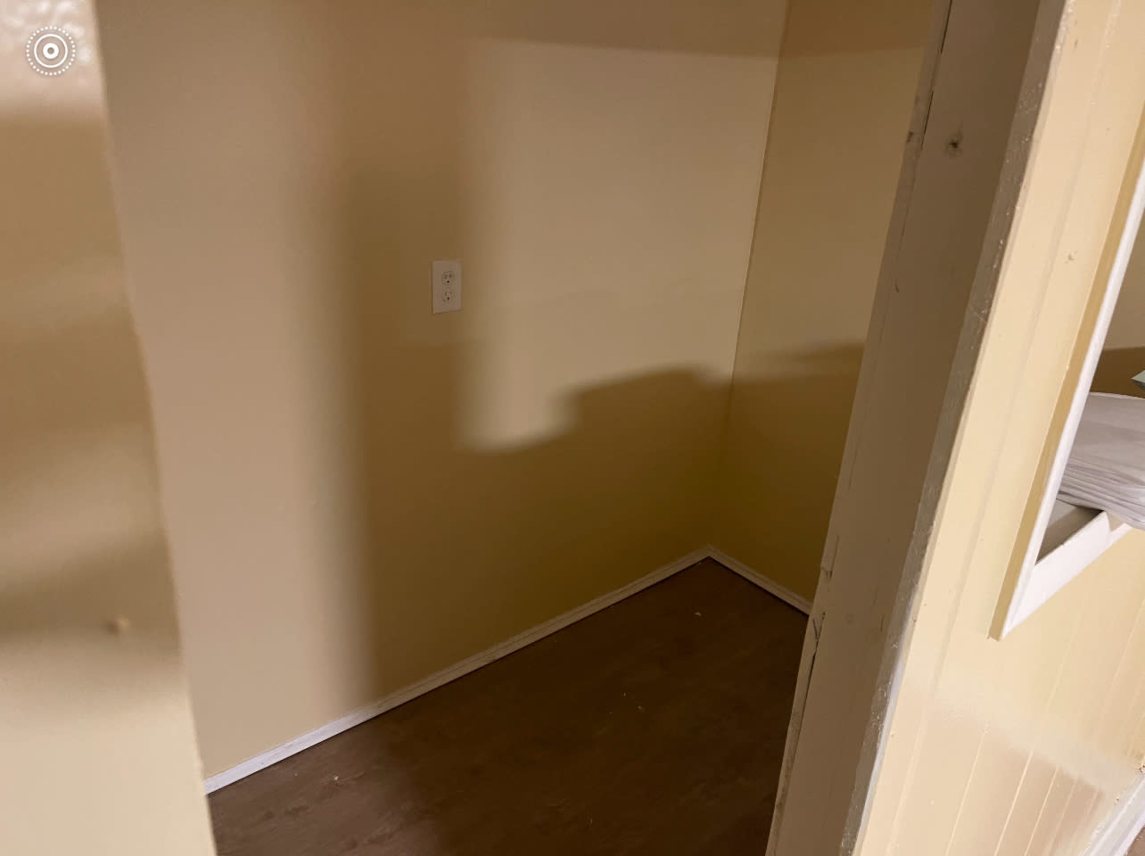 The image shows a small, empty room with beige walls and a single electrical outlet visible on one wall.