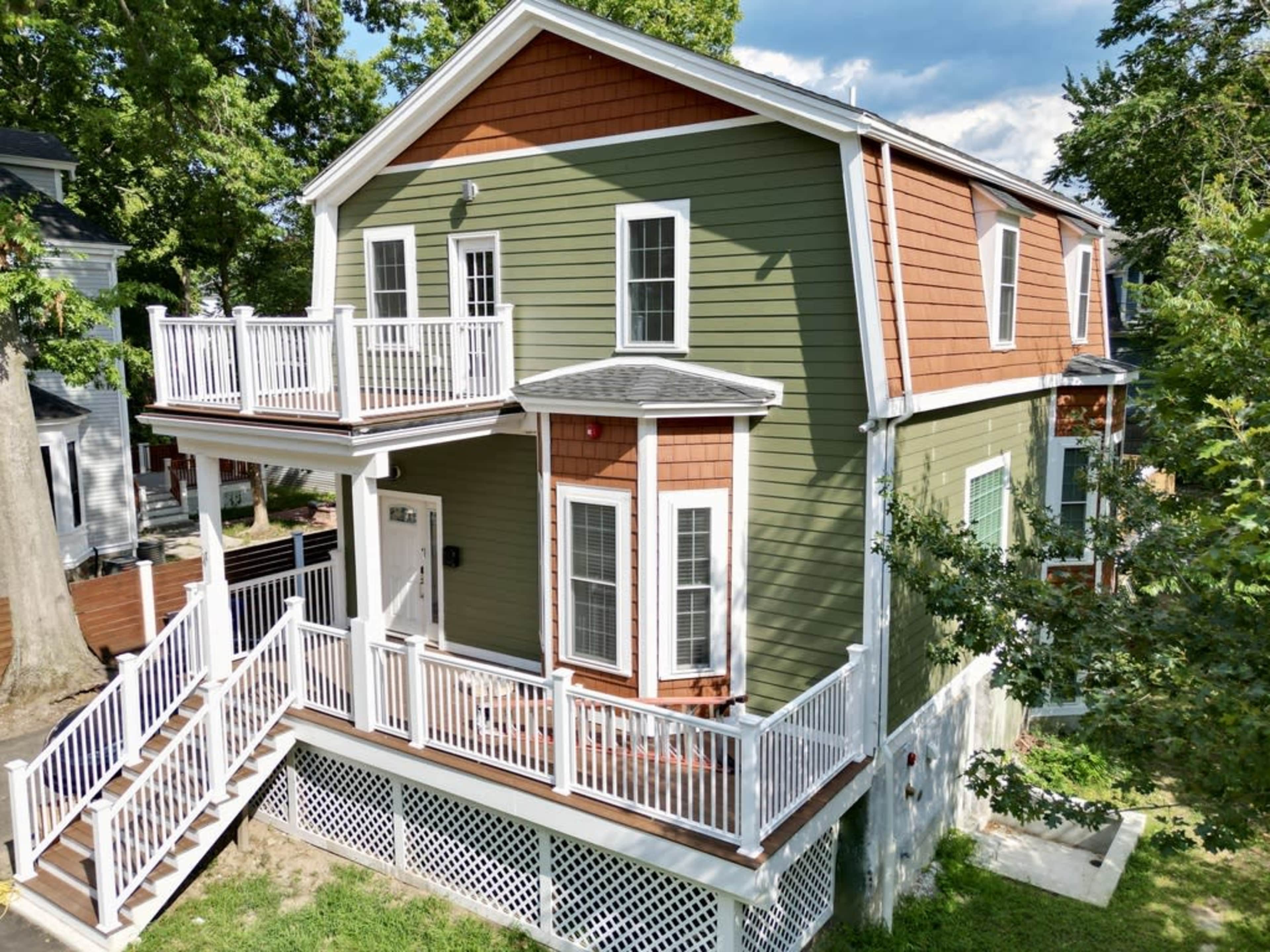 A two-story green house with a brown shingle roof, a front porch, and multiple white railings on the balconies.