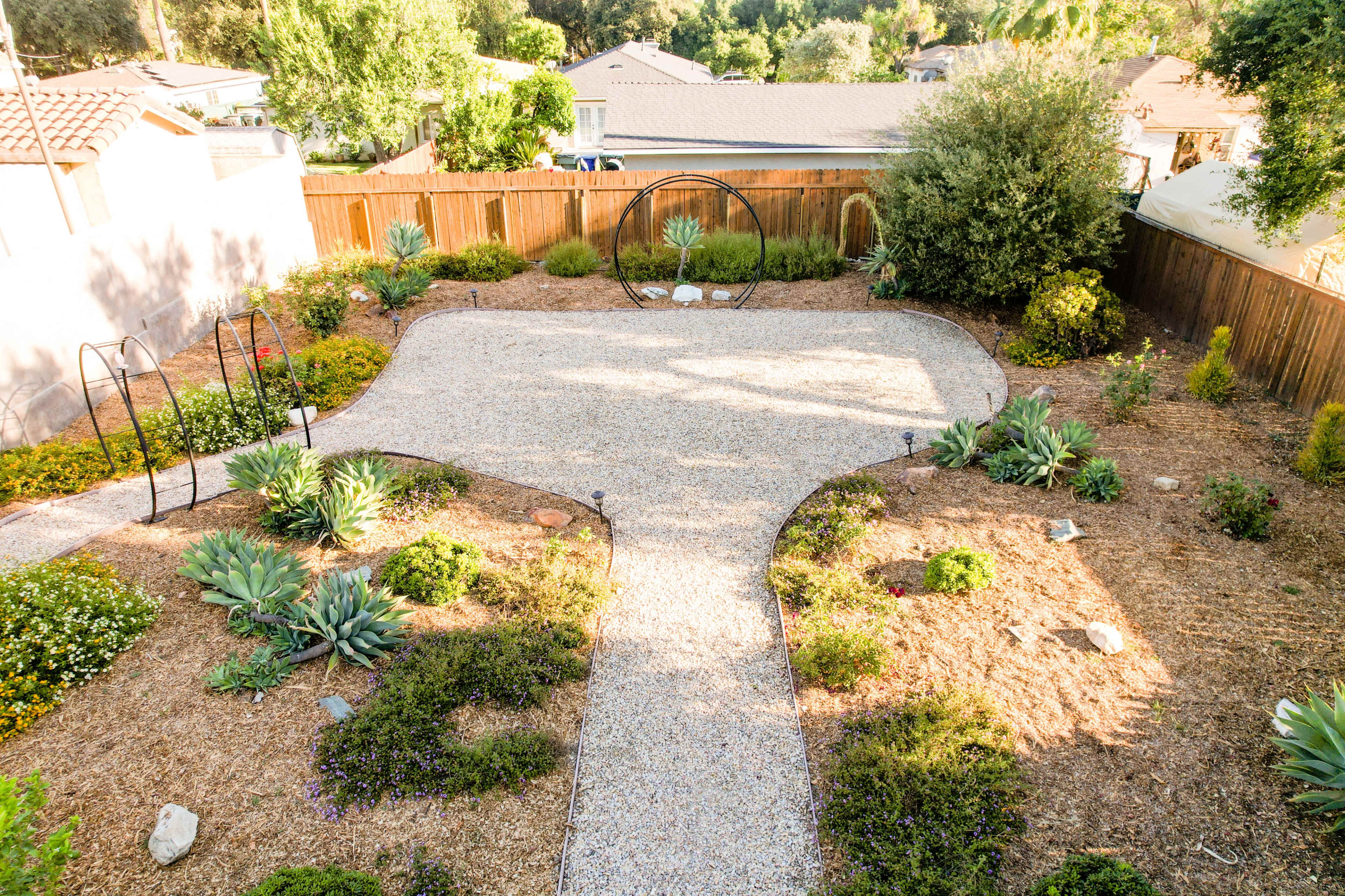 A landscaped garden features a gravel pathway outlined by various plants and shrubs, with circular structures in the background.