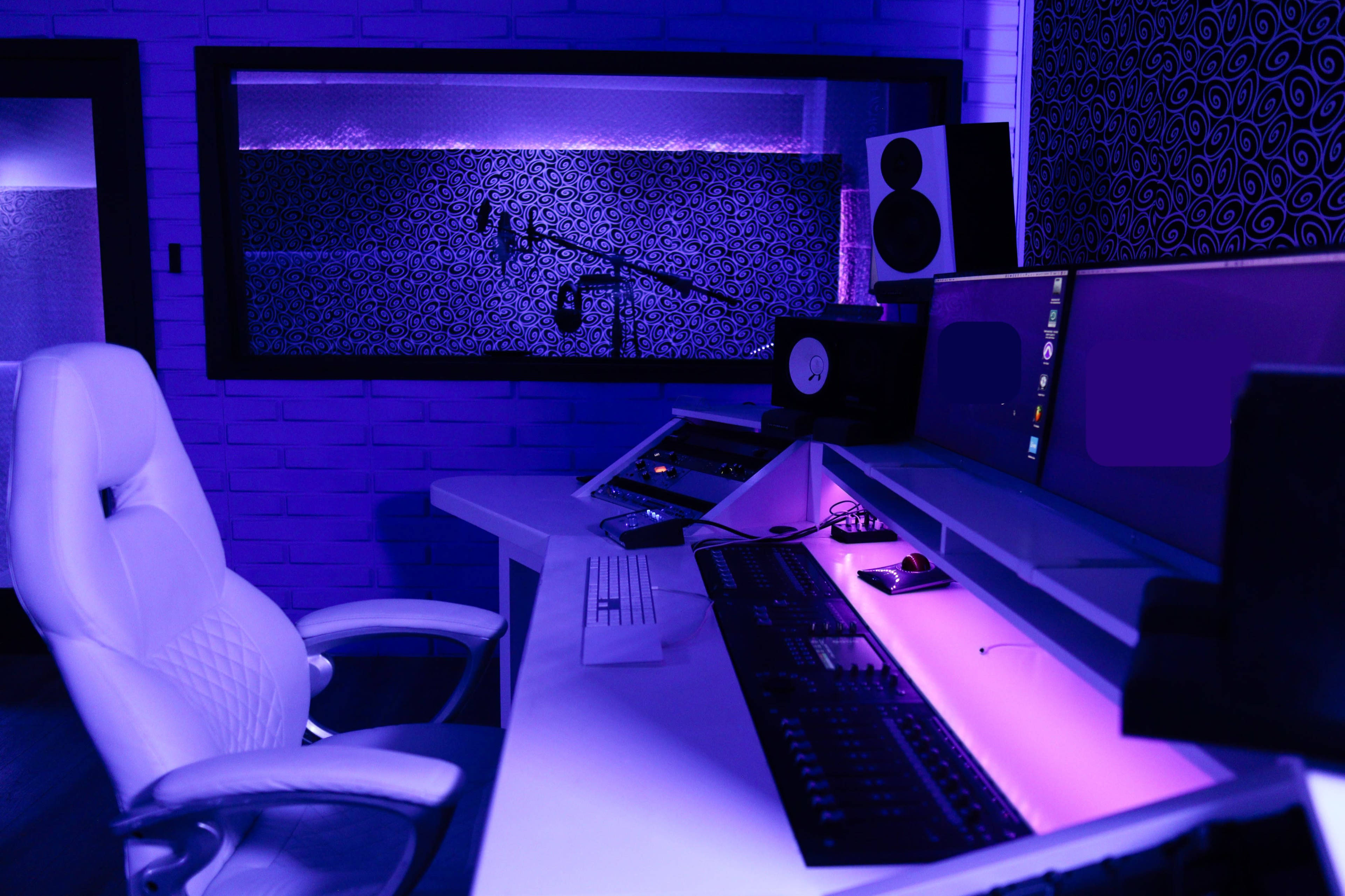 A modern recording studio with a white chair and a control desk illuminated by purple lighting.