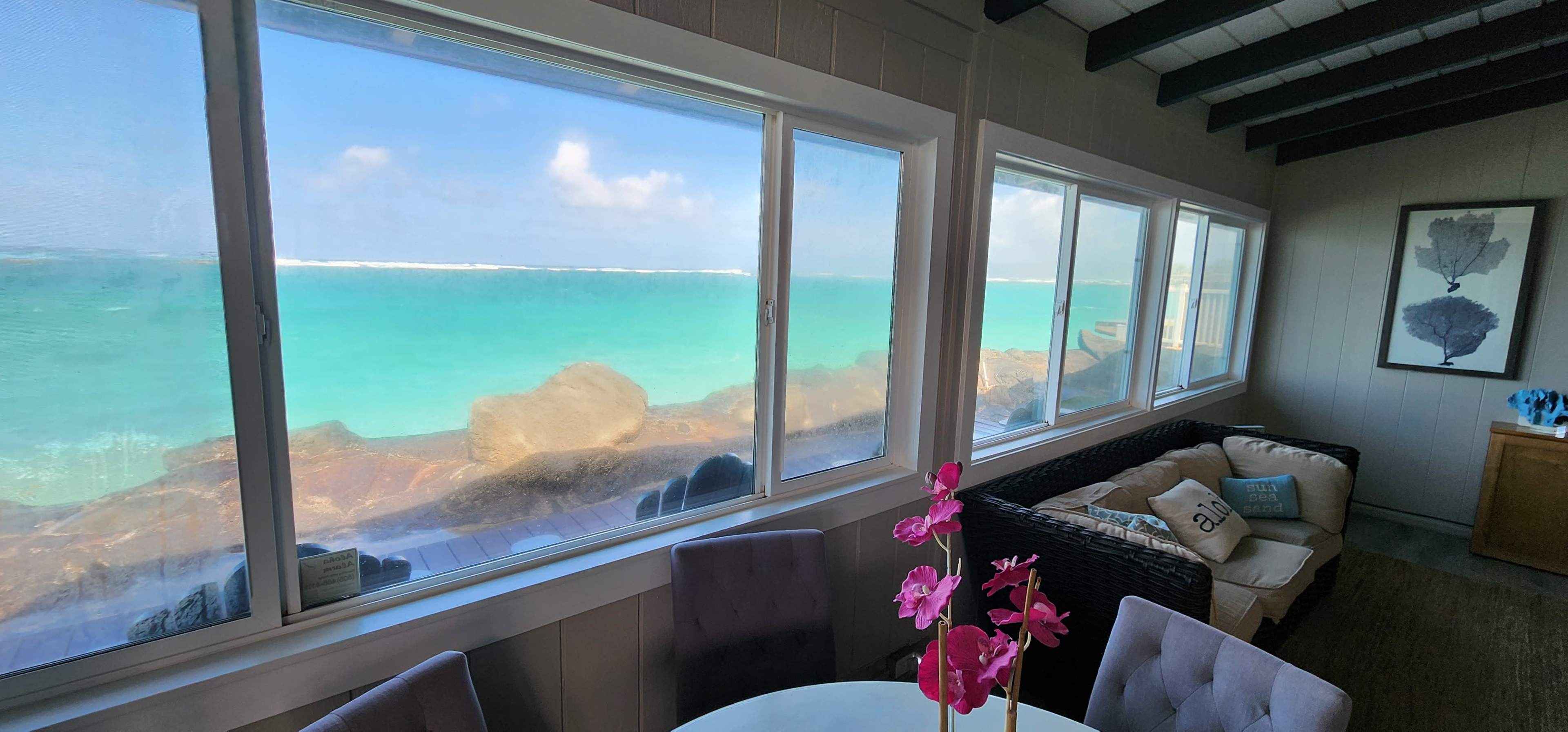 The image shows a coastal room with large windows overlooking a turquoise sea and rocky shoreline.