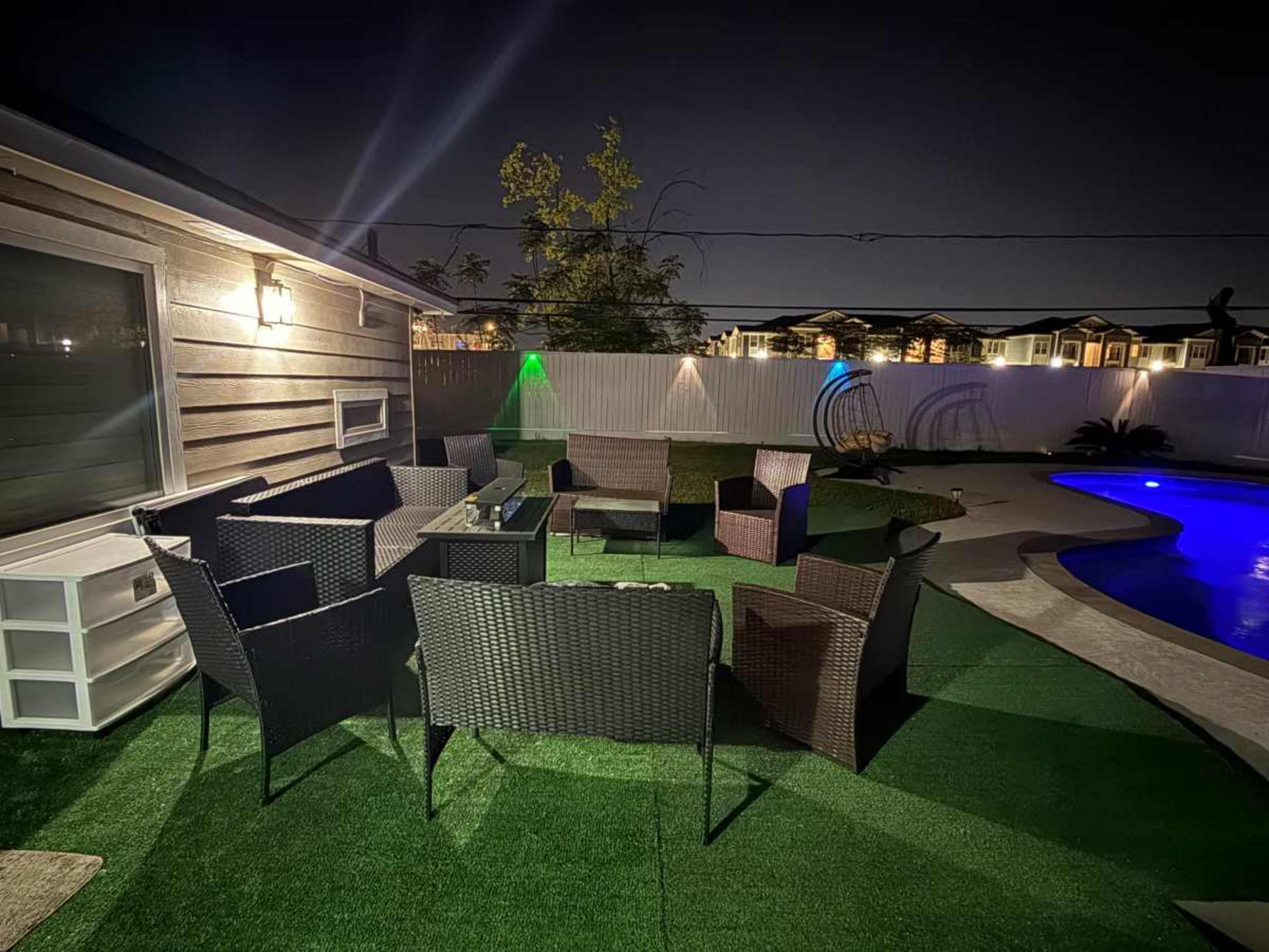 A well-lit outdoor seating area with several wicker chairs arranged around a fire pit, adjacent to a swimming pool, under a night sky.