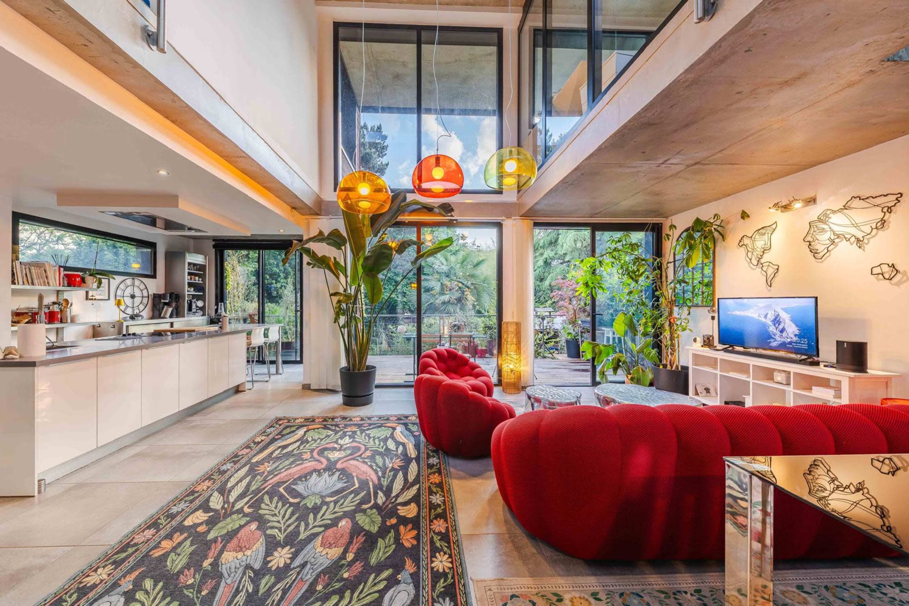A modern living room features red sofas, large windows with views of greenery, and colorful pendant lights hanging from the ceiling.