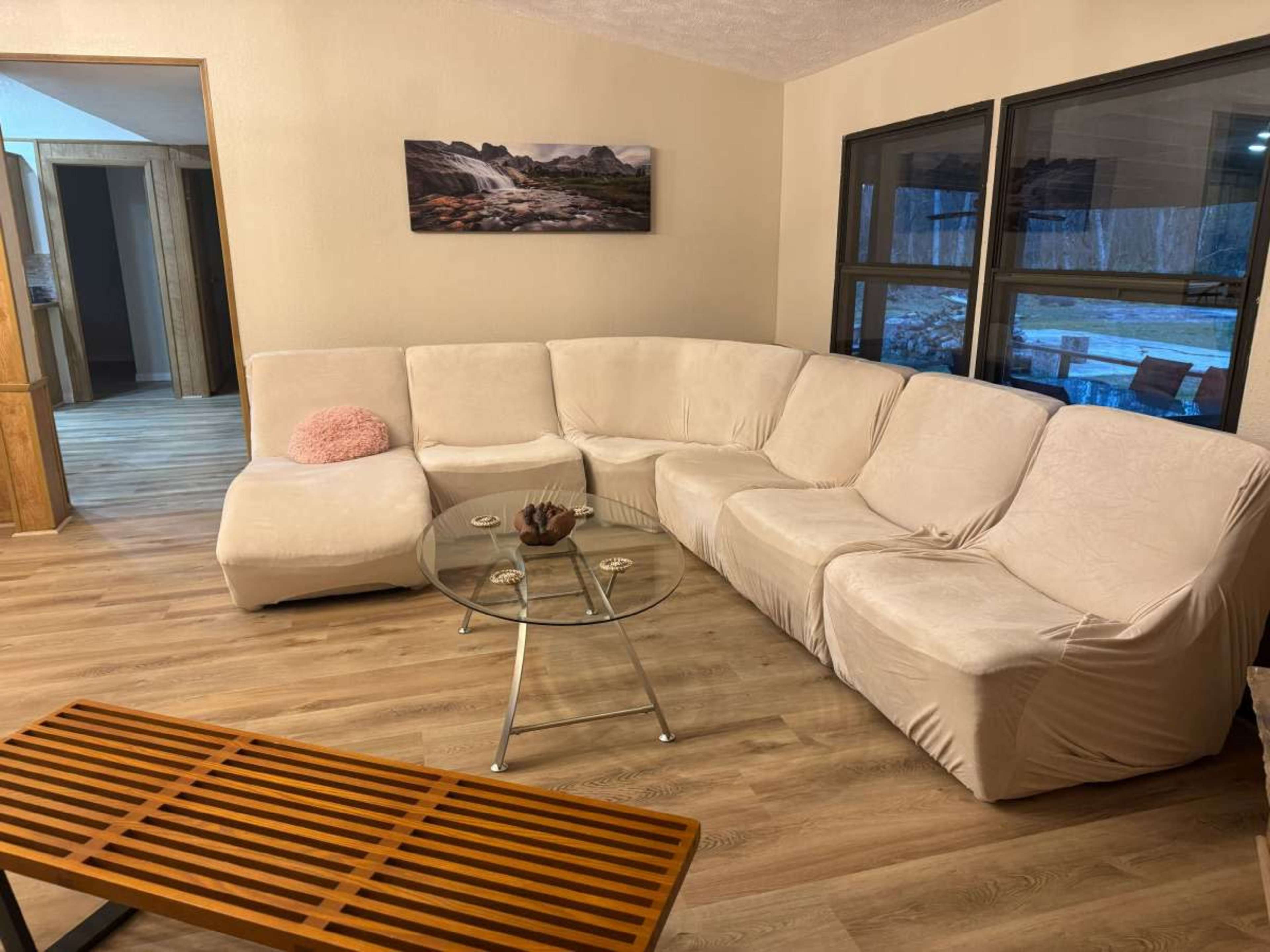 A beige sectional couch and a glass coffee table are arranged in a living room with wooden flooring and large windows.