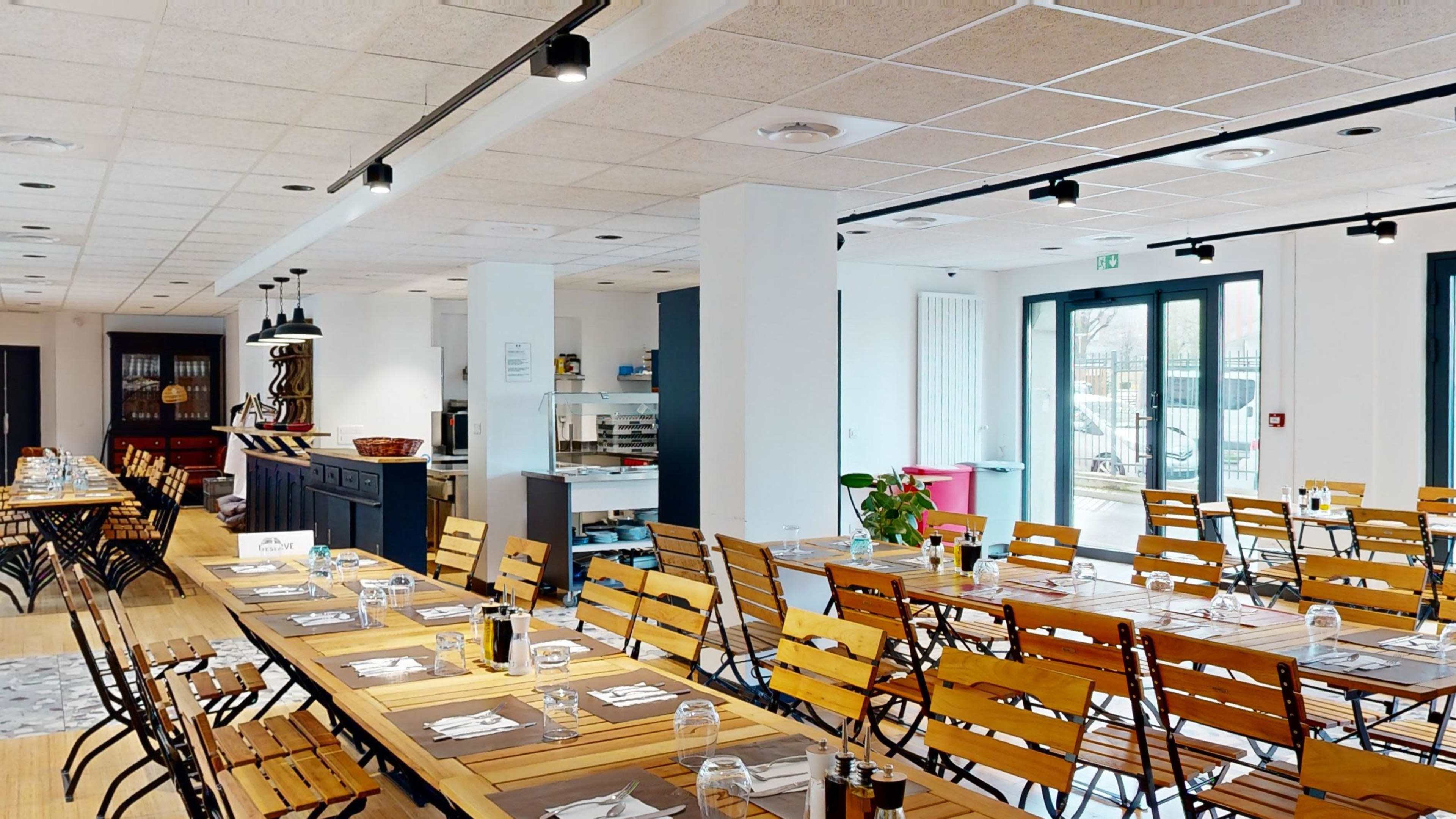 A spacious restaurant features wooden tables set with place settings and metal chairs arranged in rows, with a serving area visible in the background.