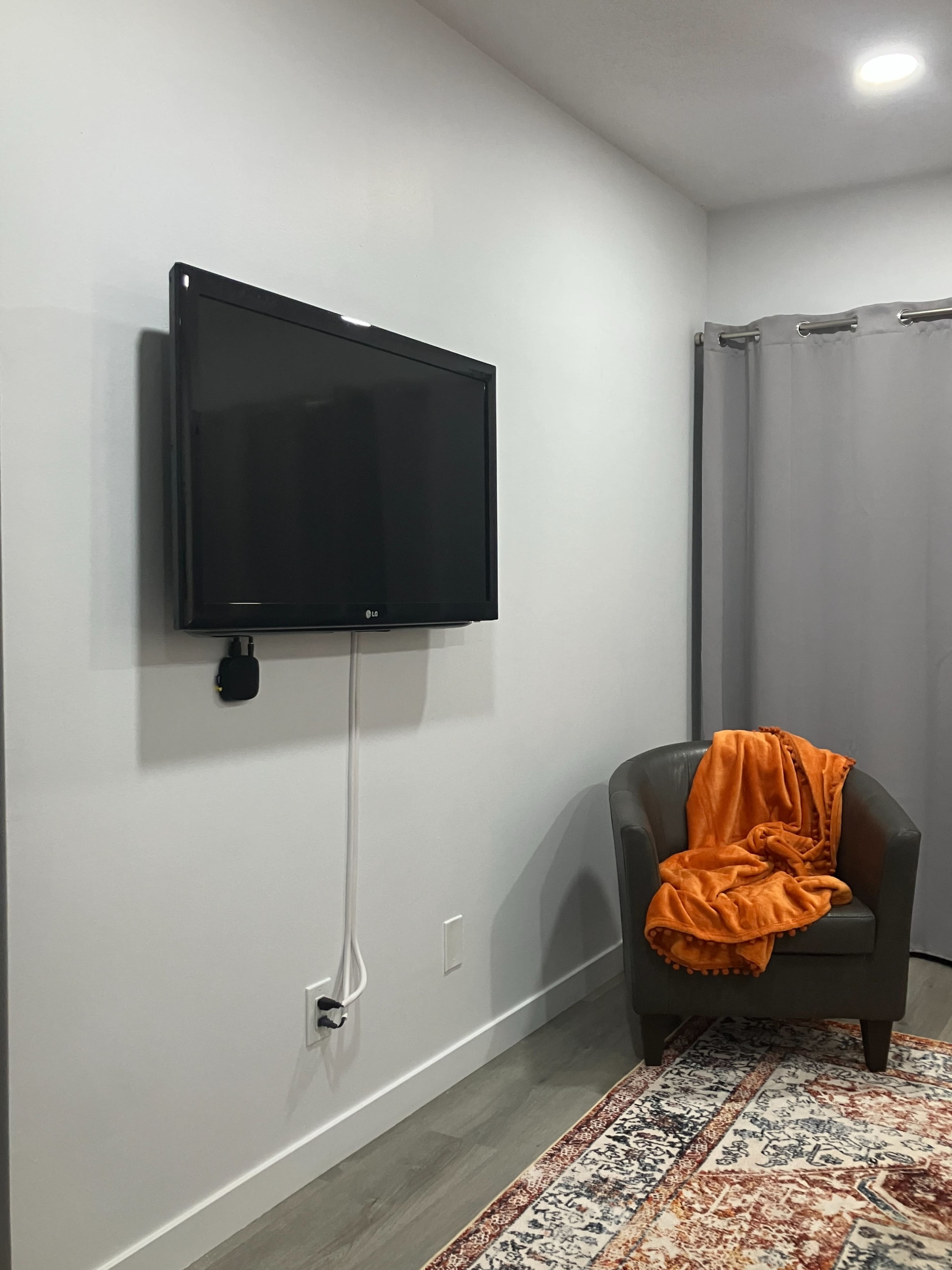 A wall-mounted television is positioned above a power outlet next to a chair draped with an orange blanket in a minimalist room.