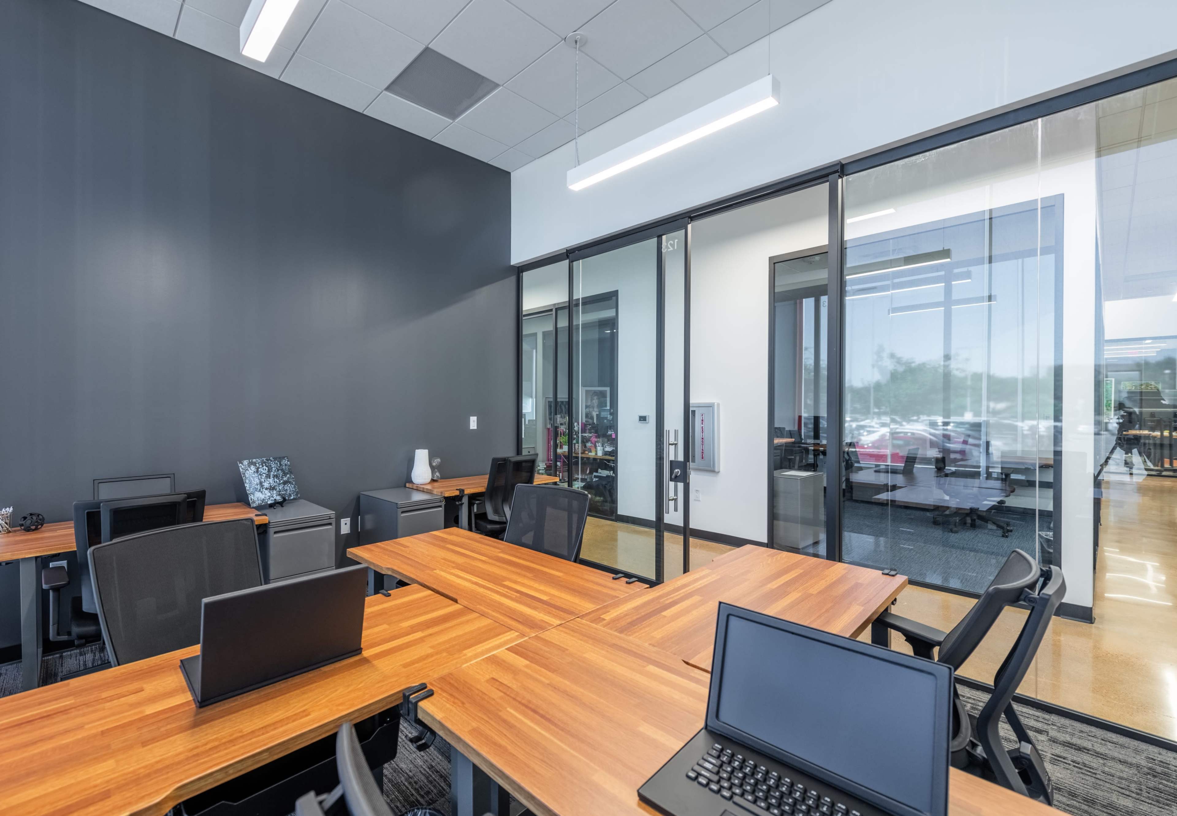 The image shows a modern office space with several wooden tables and black chairs, surrounded by glass walls and a view of an outdoor area.