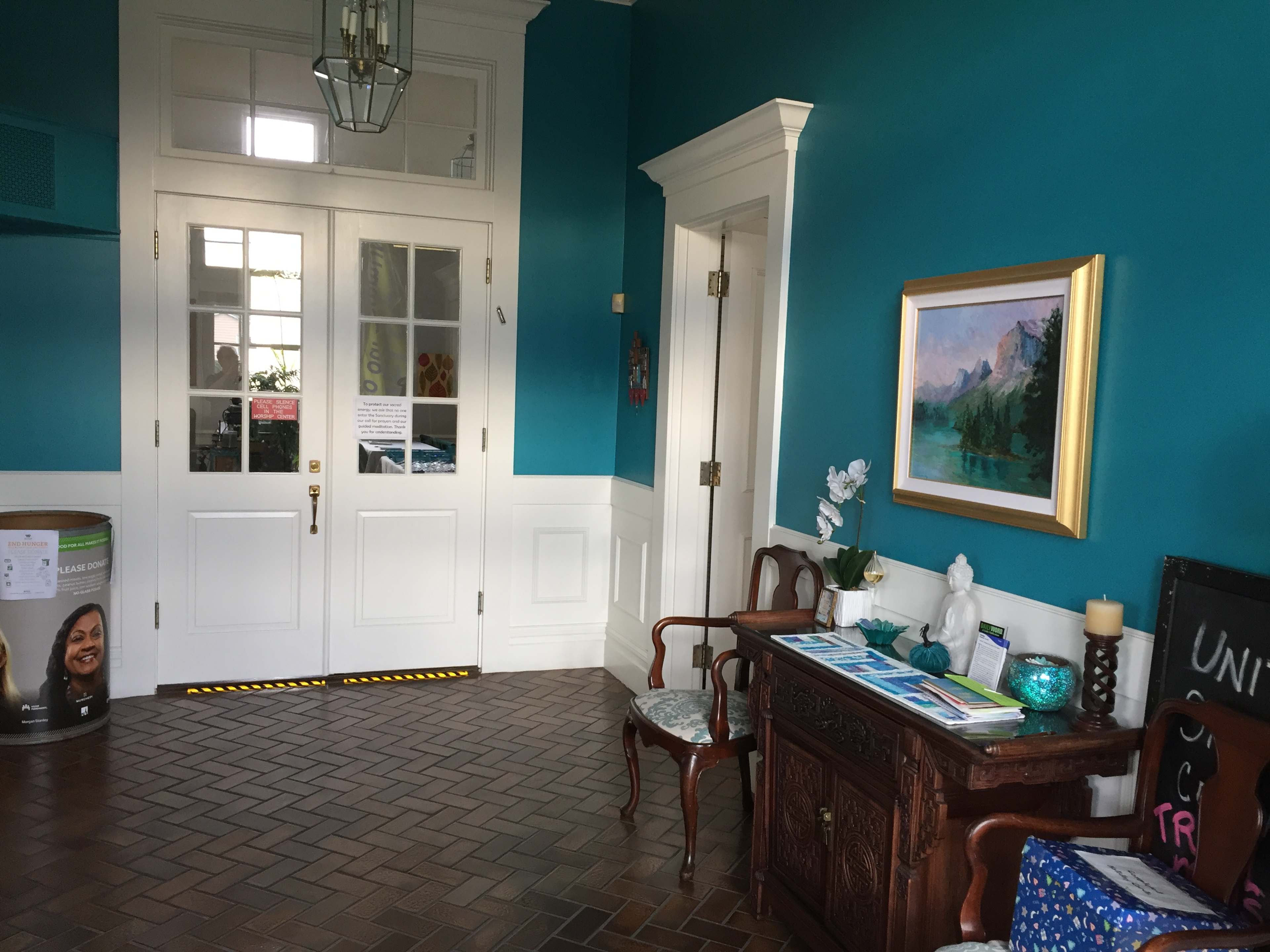 A brightly colored room with teal walls, featuring a wooden console table, a framed landscape painting, and a pair of double doors leading to another space.