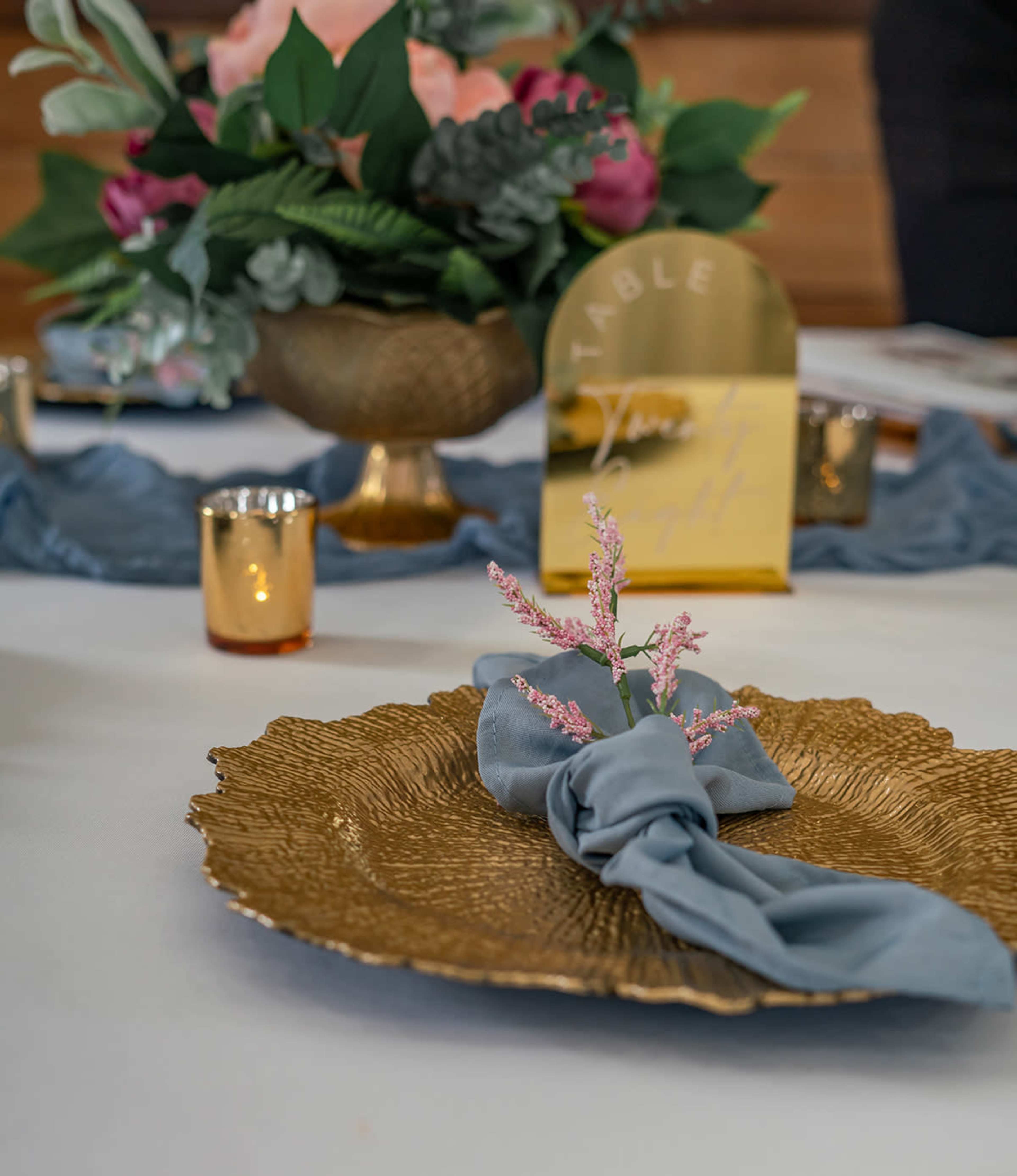A decorative table setting features a gold plate with a blue napkin and a pink floral accent, alongside a bouquet and golden votive holders.