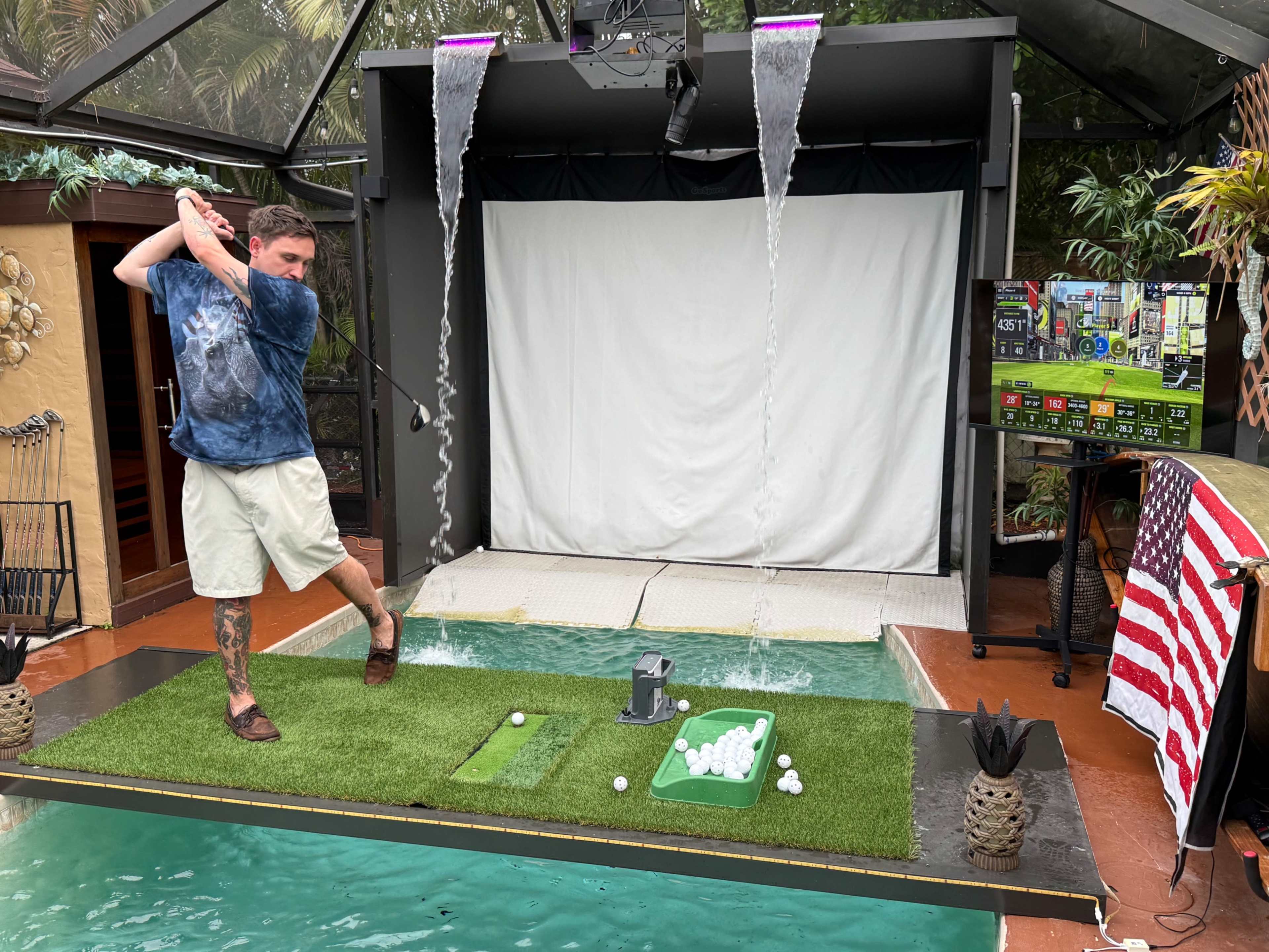 A man is swinging a golf club in an indoor setup with a hitting area, a projector screen, and an artificial putting green beside a swimming pool.