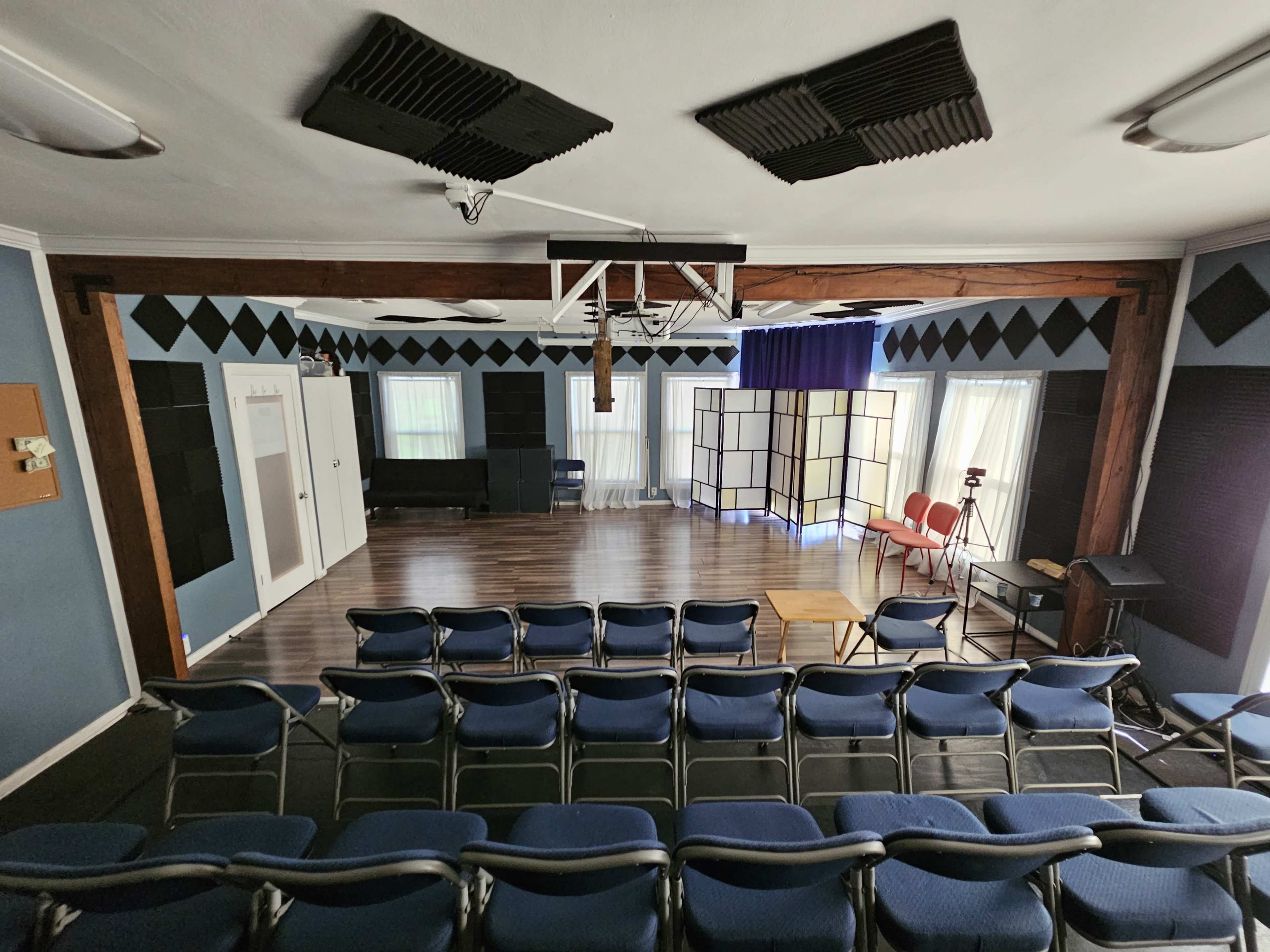The image shows a spacious auditorium with rows of blue chairs facing a stage, flanked by large windows and soundproofing elements on the walls.