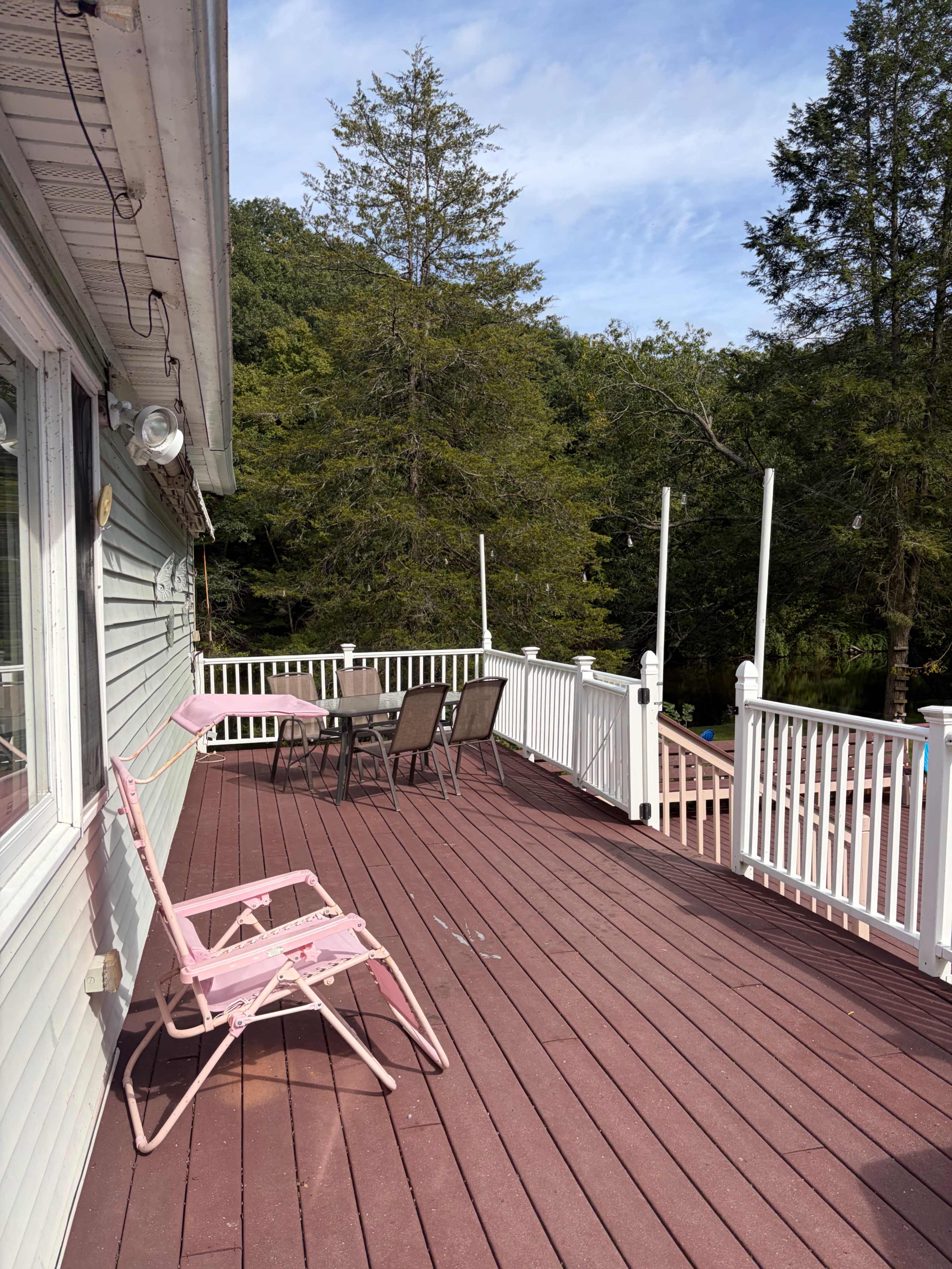 The image shows a wooden deck with a pink lounge chair and a set of metal outdoor chairs and a table, surrounded by trees and a clear blue sky.