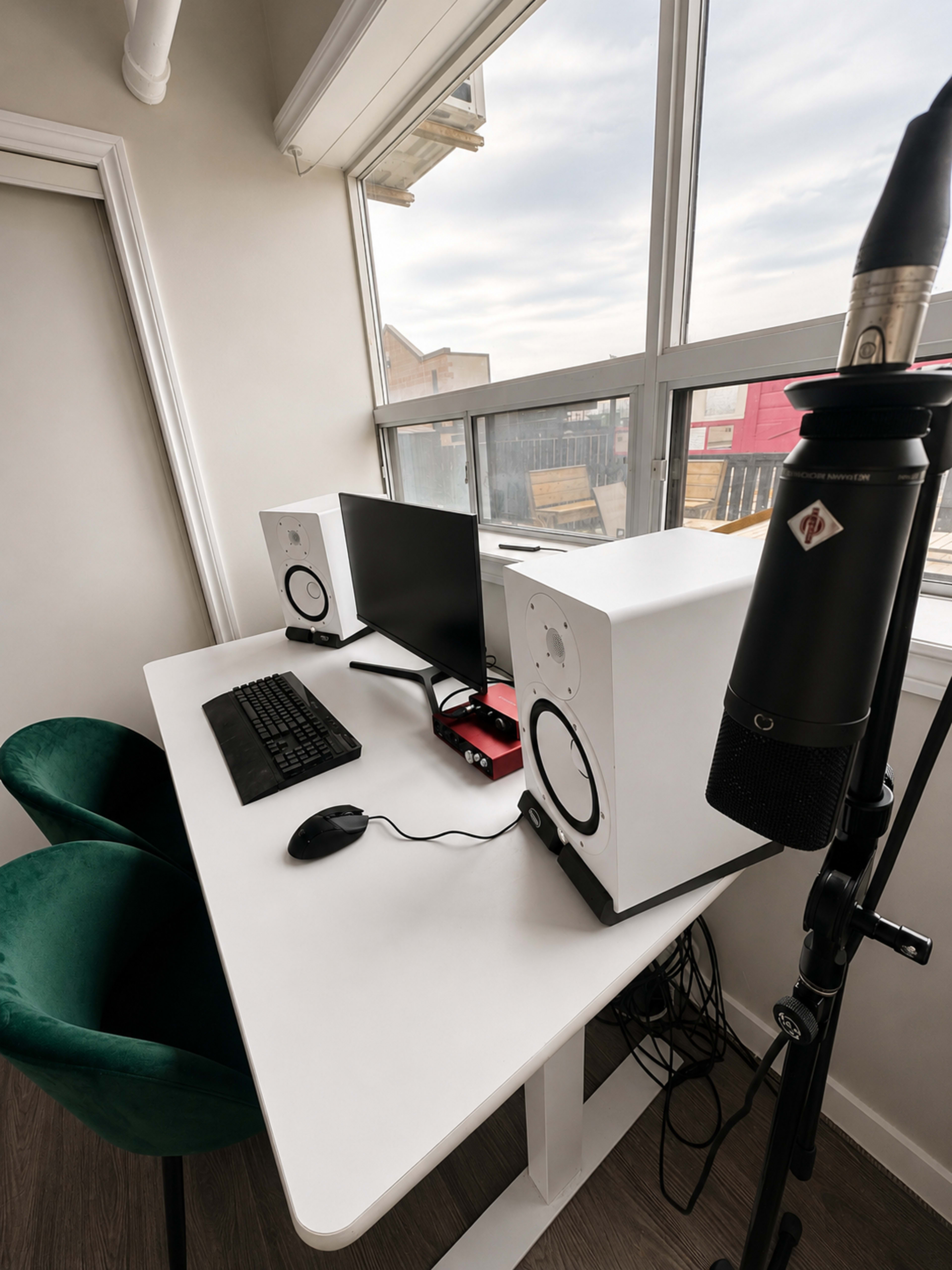 The image shows a modern workspace with a desk featuring a computer monitor, keyboard, mouse, and two speakers, along with a microphone on a stand and a green chair nearby, positioned near a large window.
