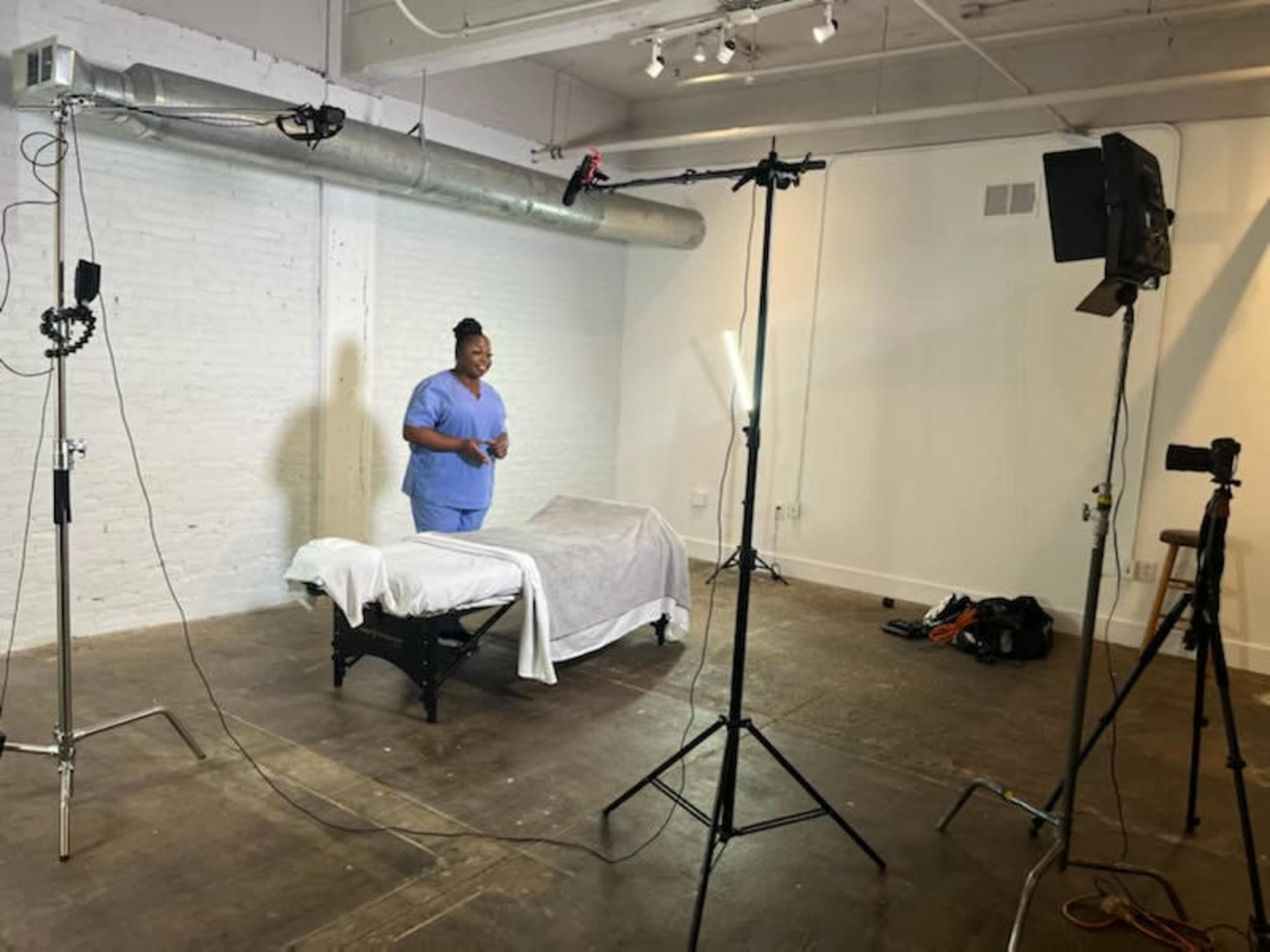 A person wearing scrubs stands in a well-lit studio next to a covered bed, surrounded by photography lighting equipment and a white brick wall.