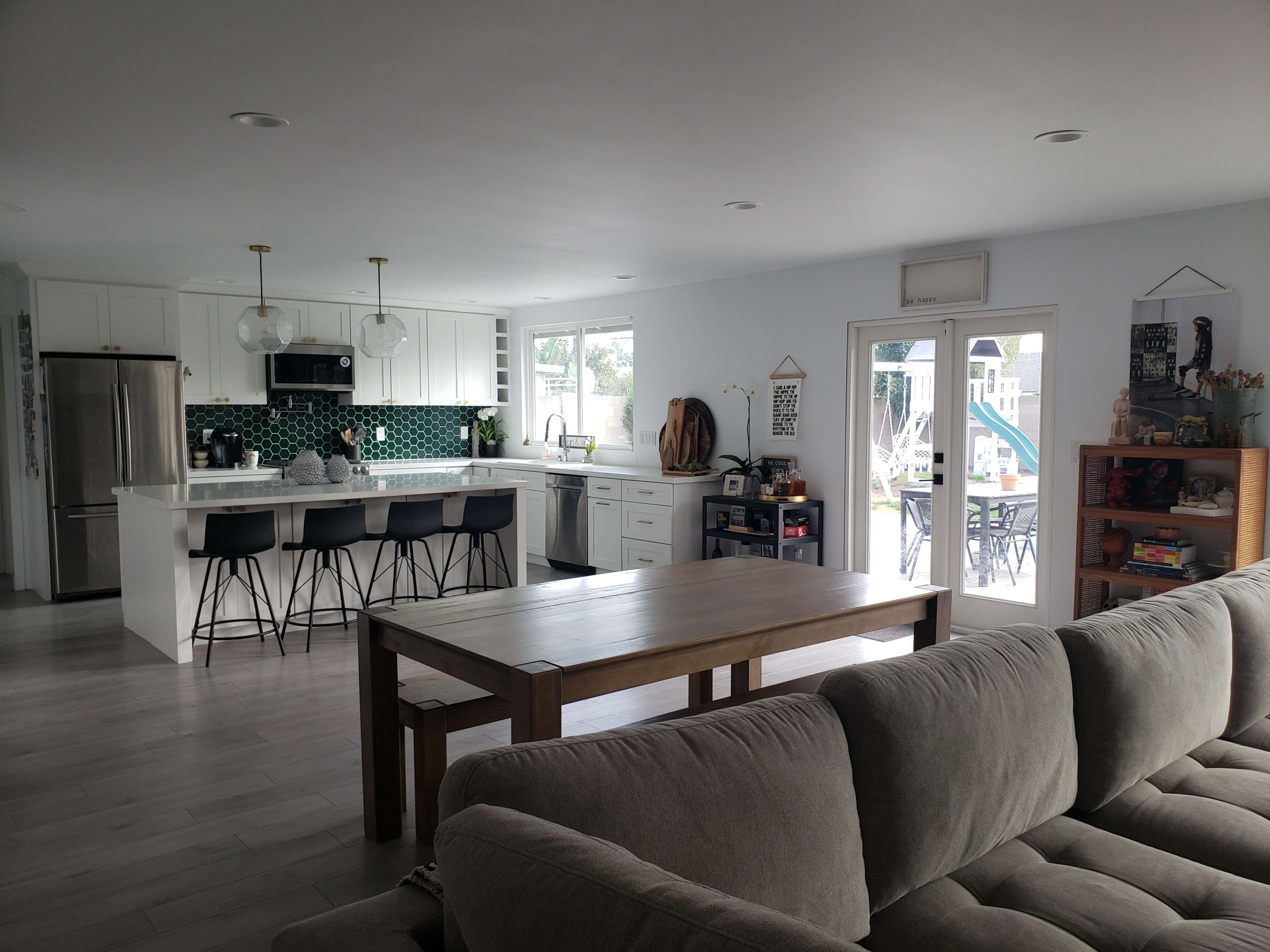 A modern open-concept kitchen and living area featuring a long wooden dining table, a large sofa, and a kitchen with white cabinets and green tile backsplash.