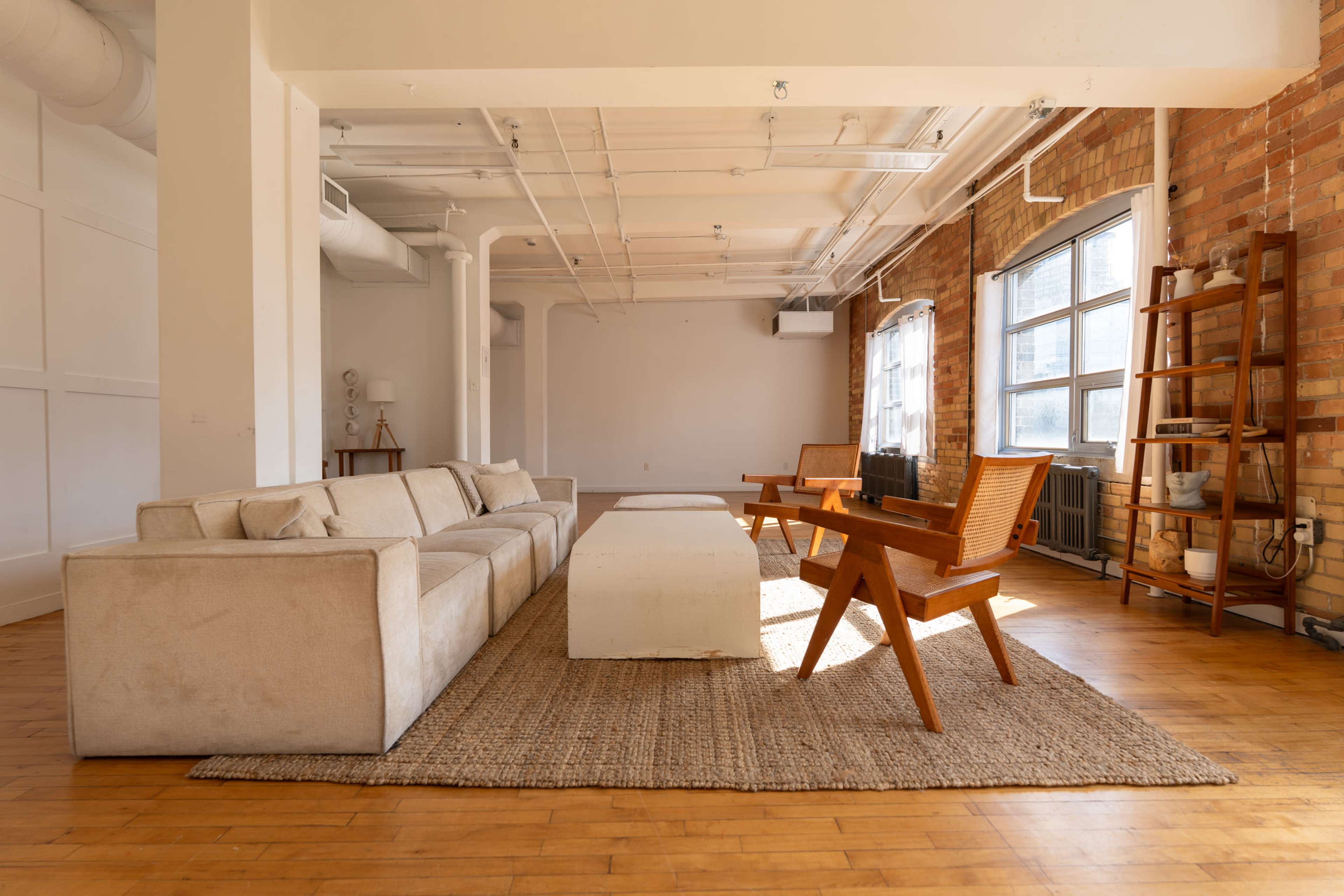 The image shows a spacious, minimalist living area with a large sectional sofa, a wooden coffee table, and two wooden chairs on a woven rug.