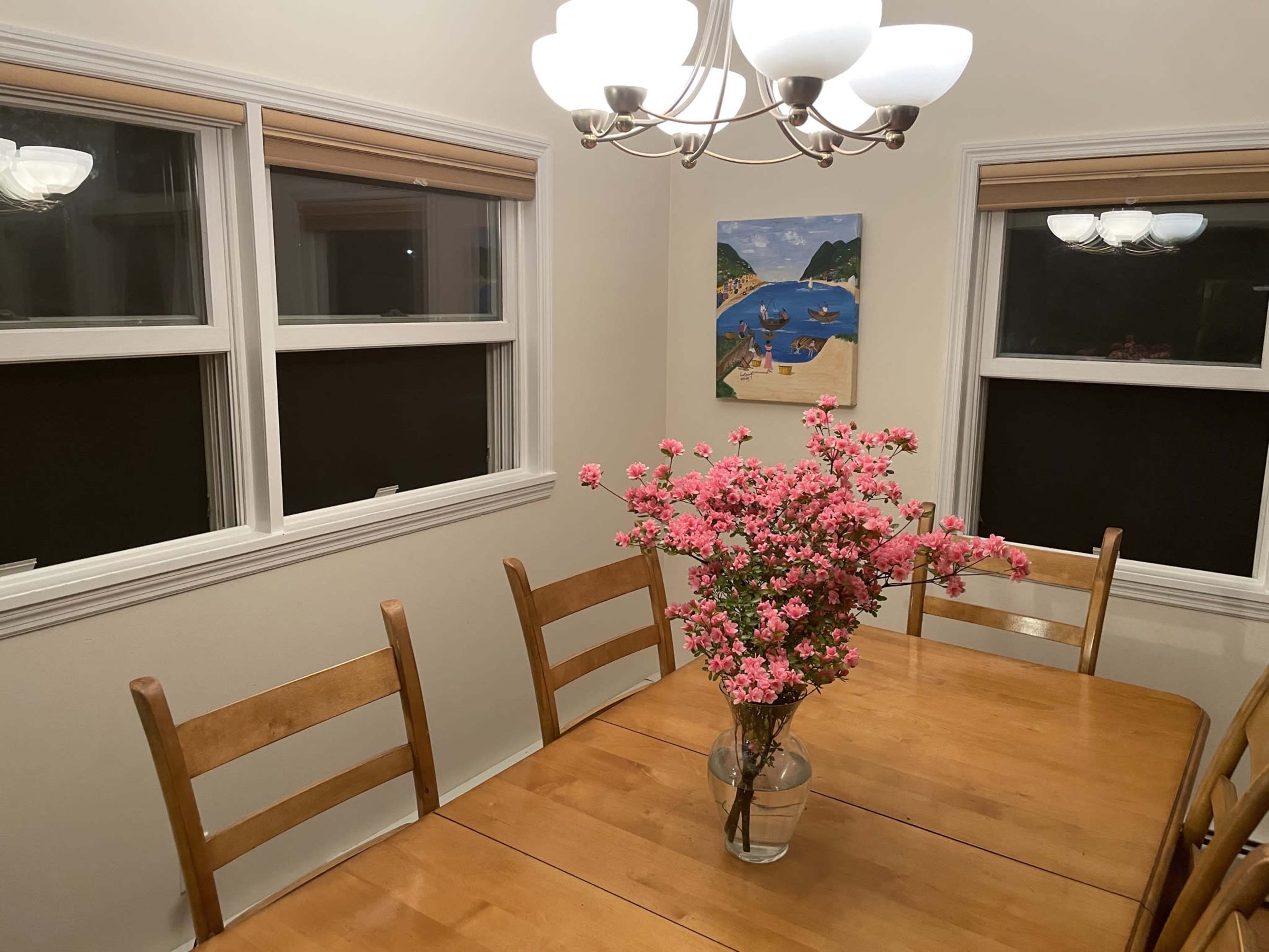 A dining area features a wooden table surrounded by chairs, with a floral arrangement in a vase and a painting on the wall.