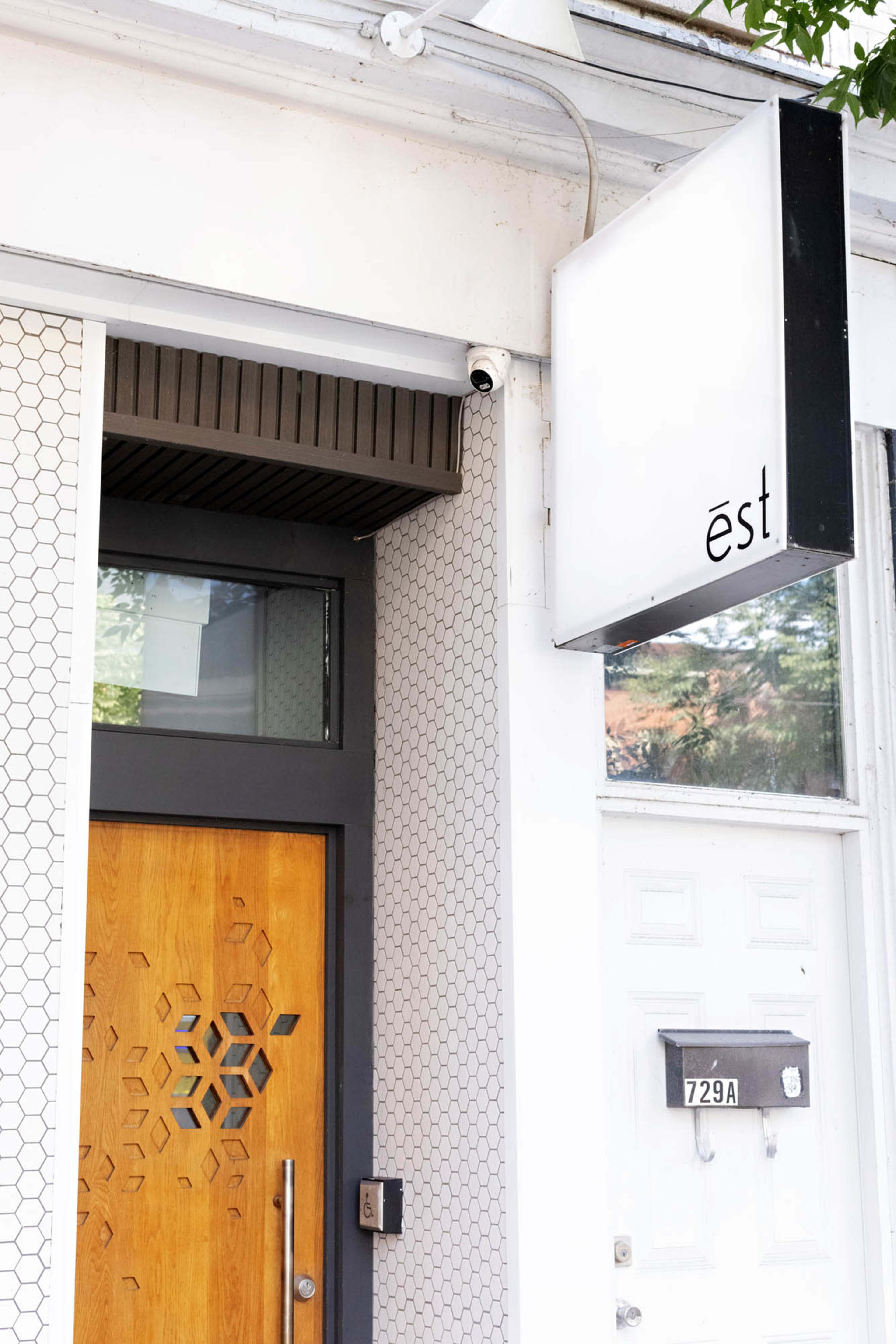 The image shows the entrance of a building featuring a wooden door with a geometric design, a hexagonal tiled wall, and a sign that reads "est."