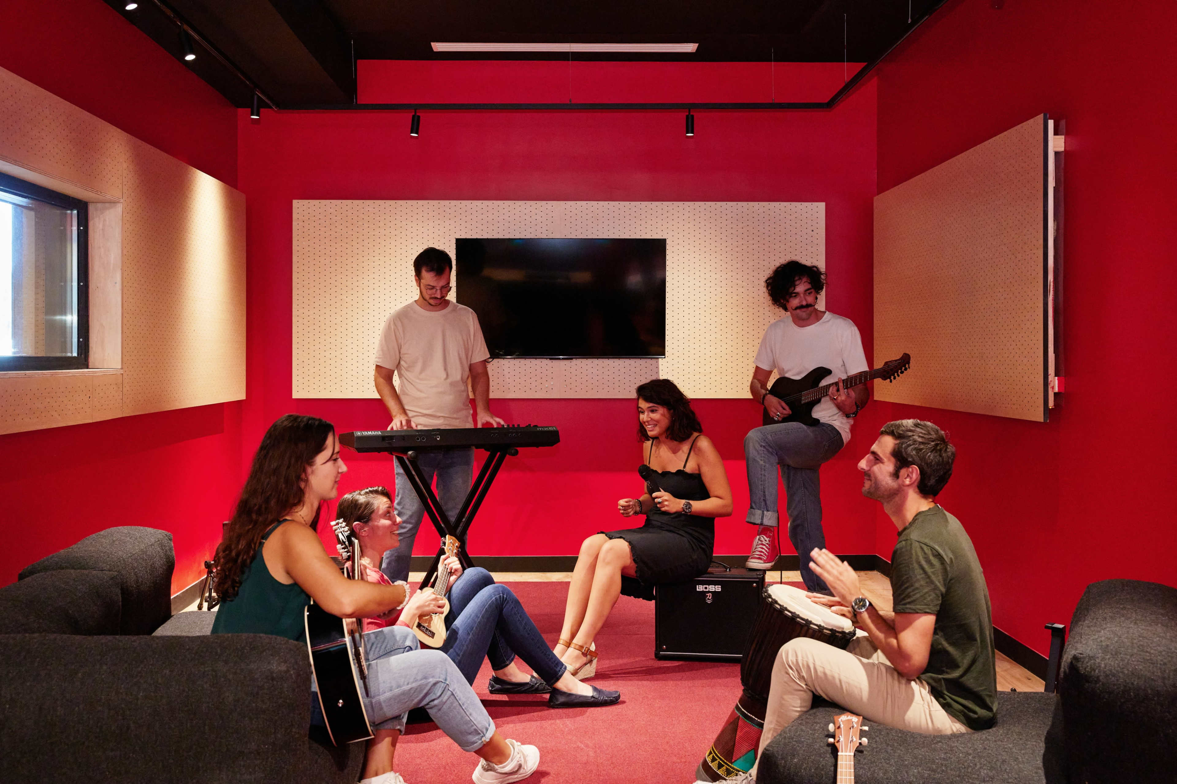 A group of six people is seated and standing in a music practice room, with one person playing a keyboard, two playing guitar and a percussion instrument, and two others participating in the session.