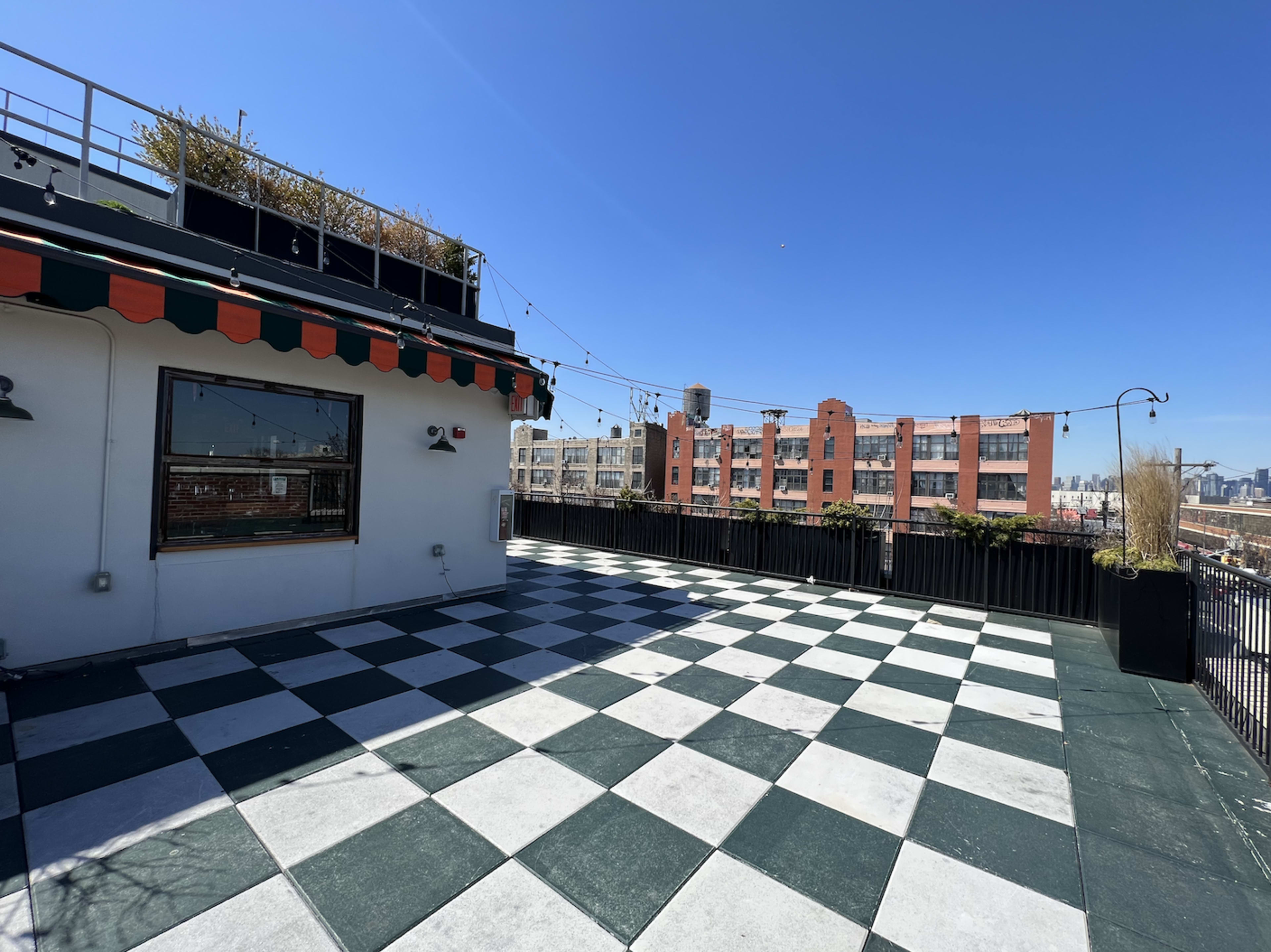 The image shows a rooftop terrace with a checkered patterned floor, featuring a clear blue sky and urban buildings in the background.